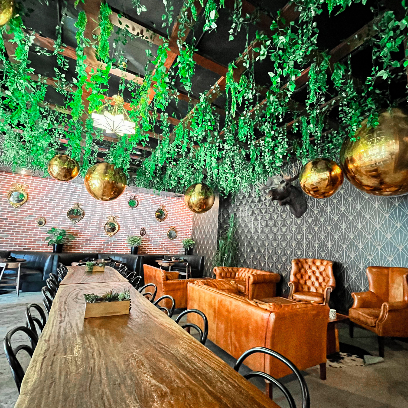 Green branches extend in layered rows down from the ceiling of a dining room at Western Proper. The tables below are wooden, with black wire chairs. Several small groupings of leather furniture are on the right side of the room. The wall to the left is exposed brick. The wall to the right features a black and gold art deco design with a black background and rows of connected gold diamond pattern lines extending upward.