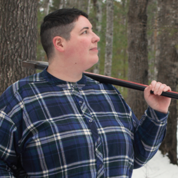 Shannon looks up and away from the camera and turns to the side. Her left hand is on her hip and her right hand is bent and holding an ax that rests also on her right shoulder. She is wearing a blue-and-white plaid flannel shirt and jeans. Part of a split log is in the foreground, a snowy forest with bare trees is behind her.