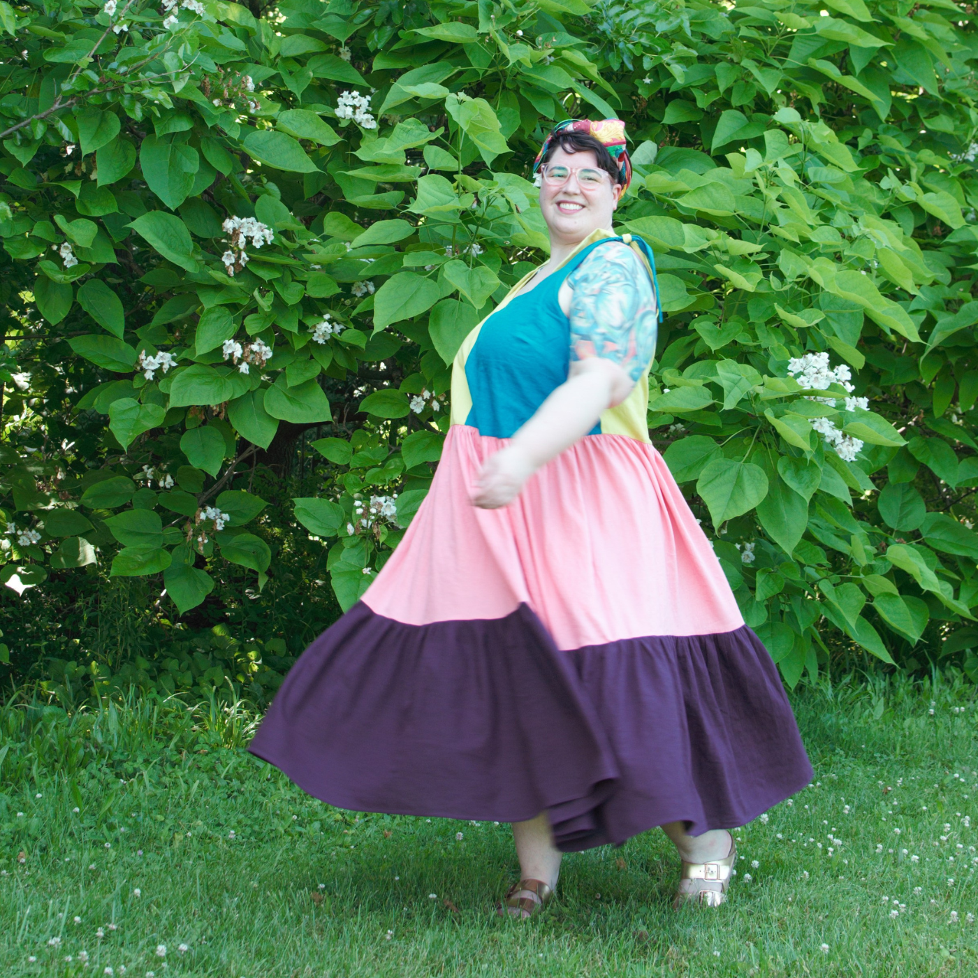 Shannon looks at the camera mid-spin in the grass. She is wearing a multi-tiered dress (a camisole vertically half yellow and blue, leading to a pink tier and then a purple tier that lands slightly above her ankles) and gold sandals and has a red, yellow and green headband. She is standing in front of a row of tall leafy bushes, some with bursts of small white flowers.