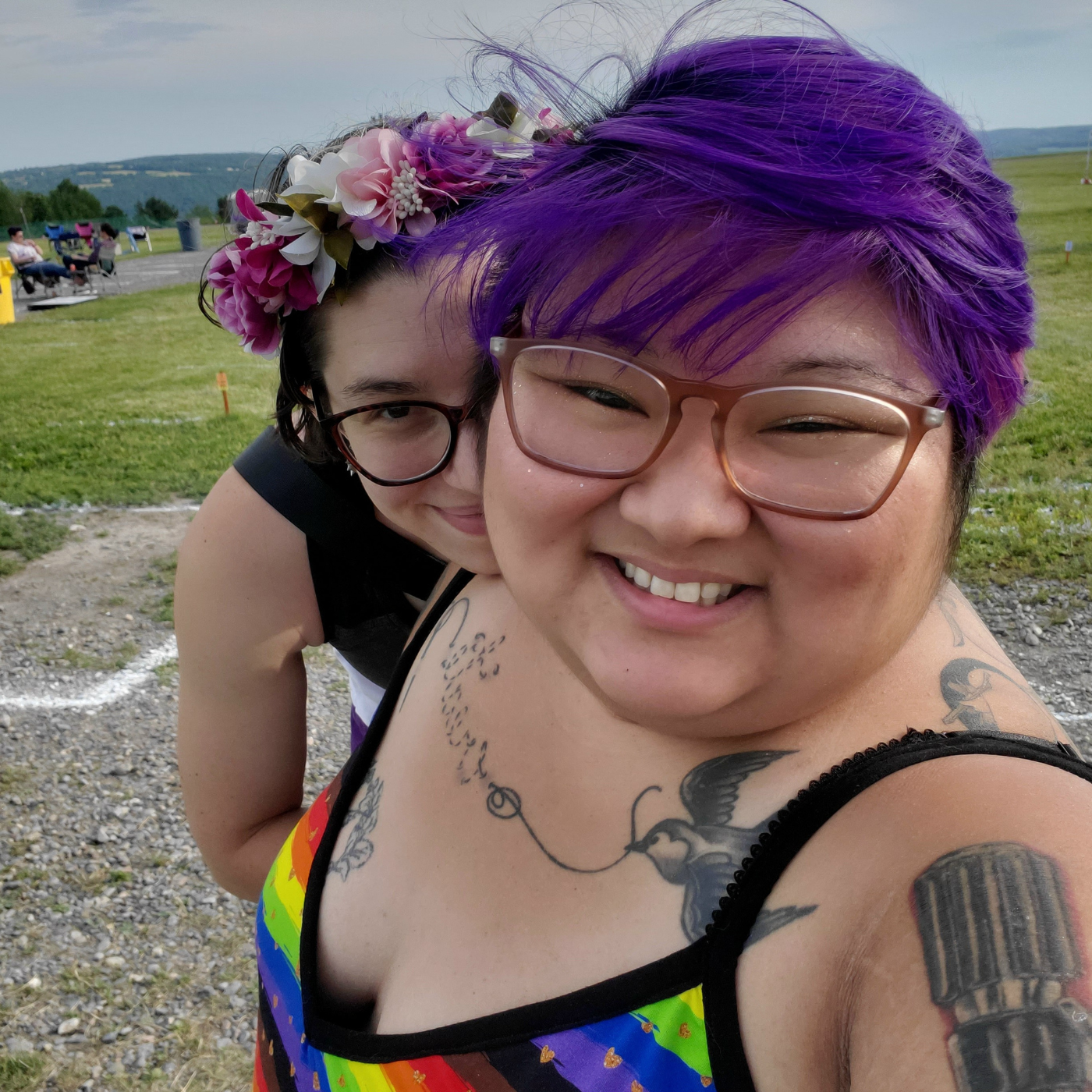 Teukie and their partner Sky look at the camera and smile. Teukie wears a tank top with black straps and a rainbow striped design with small ditsy gold hearts. Sky is wearing a flower crown and a black tank top.