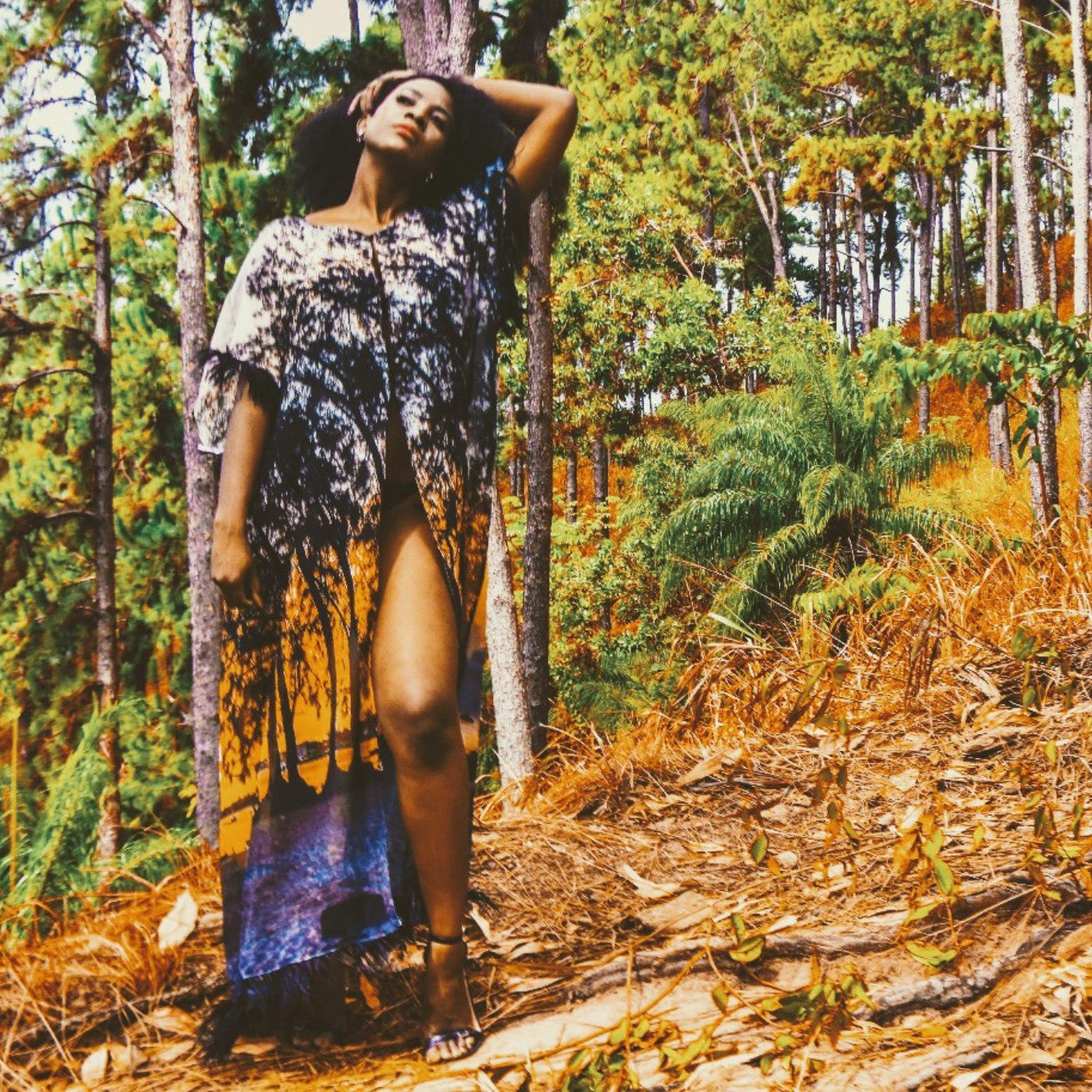 A woman stands in a grove of trees with her left arm up held behind her head. She is wearing a caftan with a print that shows a beach scene at twilight, with an orange sky, trees, sand and a gray sky.