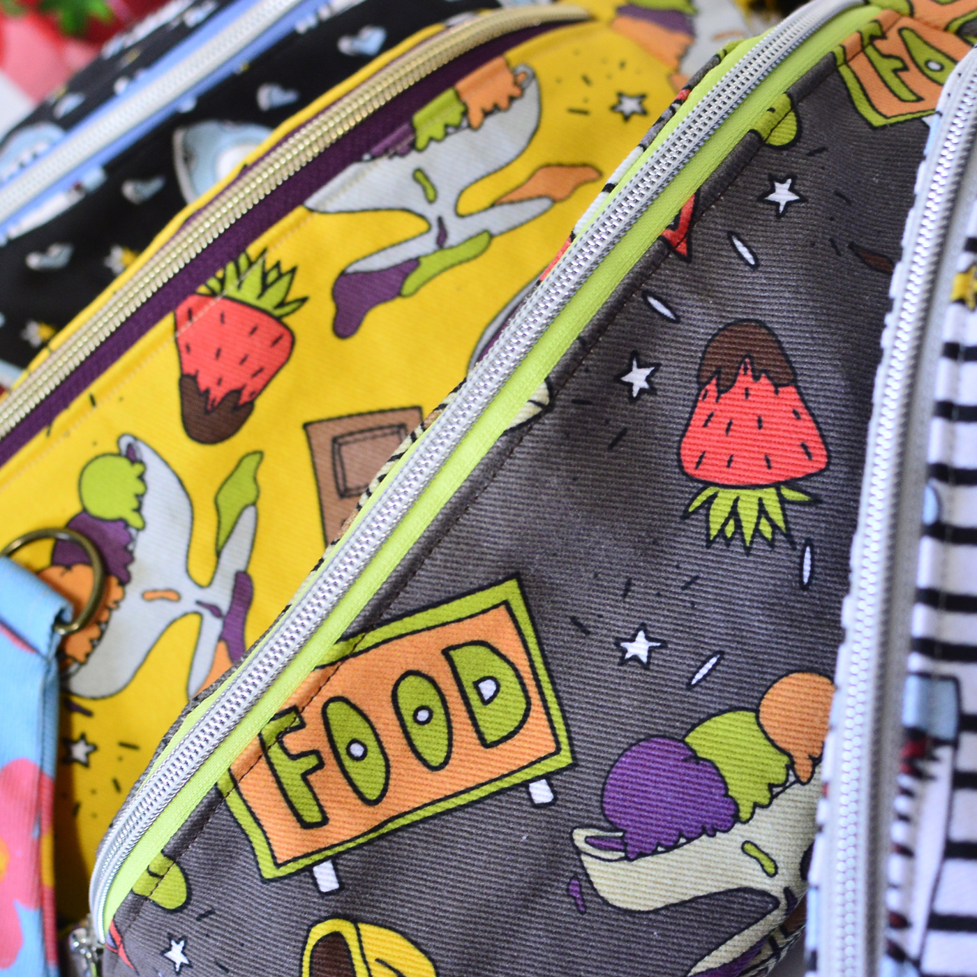 Close up of four small zipper pouches. The bag on the far left has a black background and light blue sharks with their mouths open, surrounded by small light blue hearts. The next two bags have designs of chocolate covered strawberries, ice cream sundae bowls each with three scoops of ice cream, one orange, one purple, one green, squares of chocolate and the word FOOD spelled out as if on a road sign, one bag has a yellow background and the other bag has a gray background. The bag on the bottom right has the same open-mouthed shark design with small yellow stars and a black-and-white striped background.