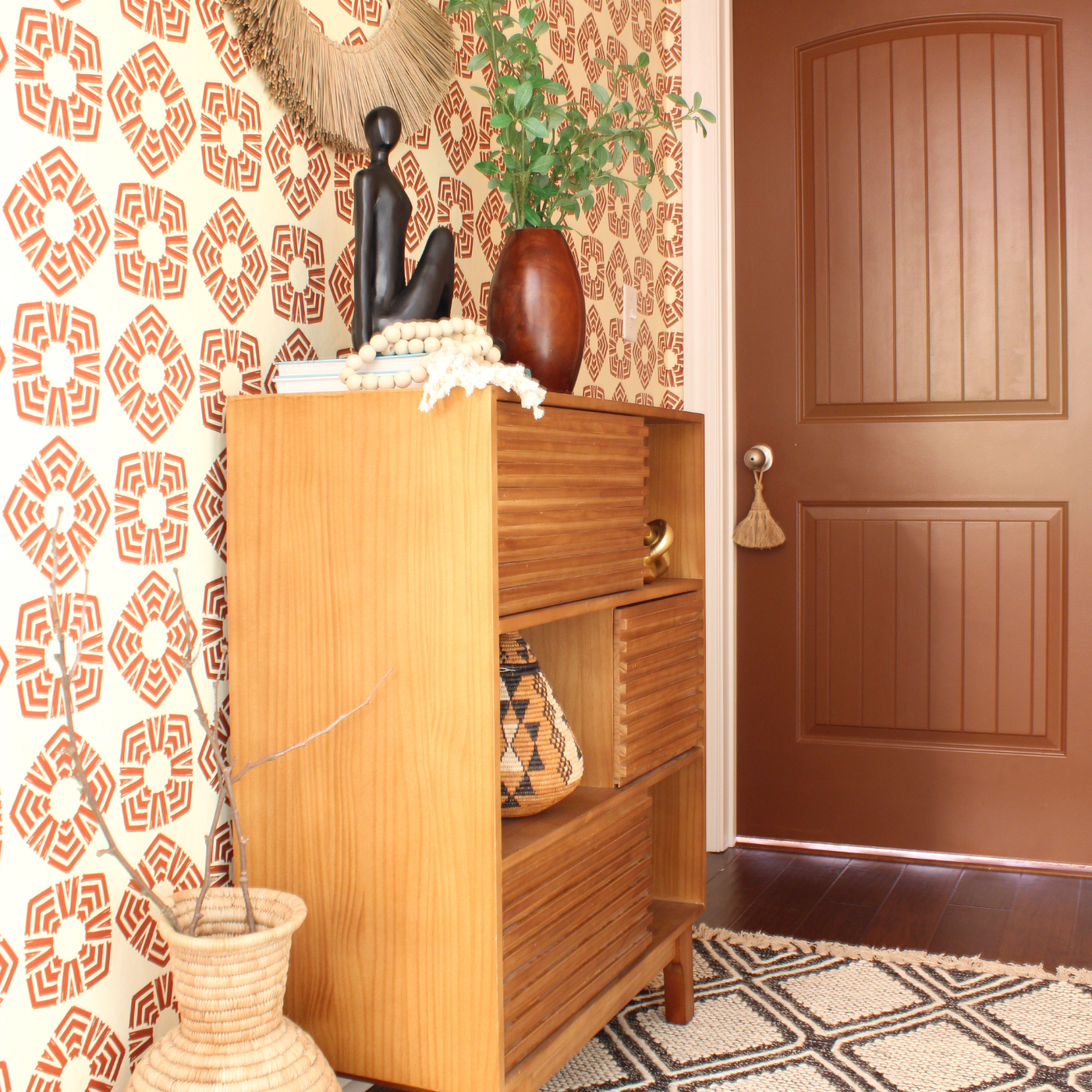 Image of a corner of a room with a brown wooden bookshelf with plants and a sculpture on top. A cream rug with brown geometric shapes is in front of the bookshelf. A wallpaper design with a cream background and repeating rows of large light brown squares is on the wall behind the bookshelf.