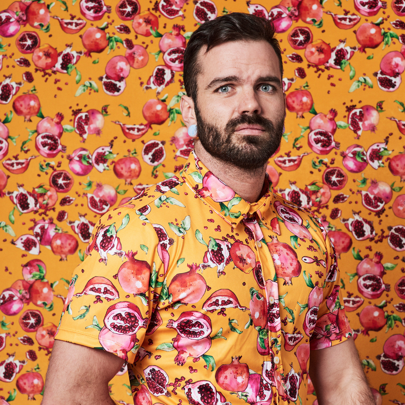 Anthony stands in front of a wall with his right hand behind his back and wears a shirt with an orange background and a design with pink and red pomegranates with green leaves. The wallpaper on the wall behind him is the same design as his shirt.