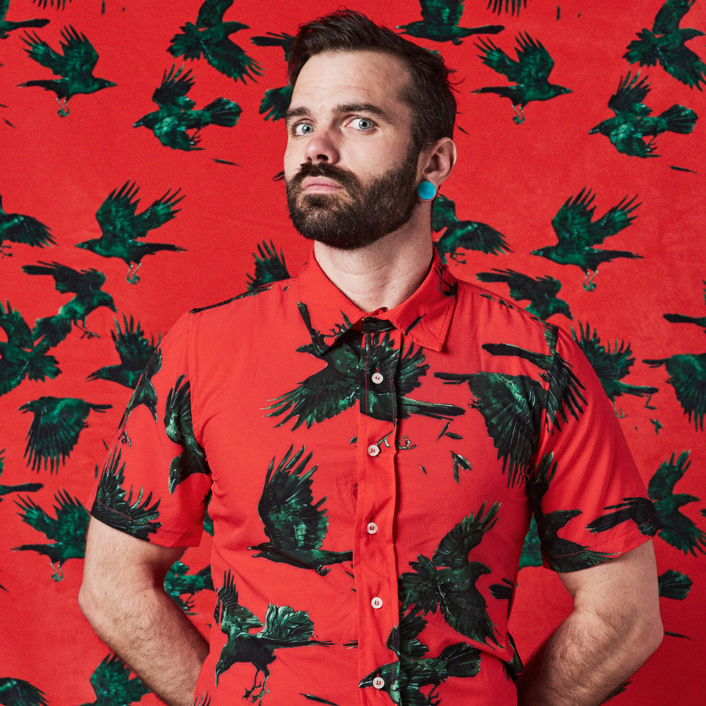 Anthony stands in front of a wall with his hands behind his back and wears a shirt with a red background and a design with green and black ravens. The wallpaper on the wall behind him is the same design as his shirt.