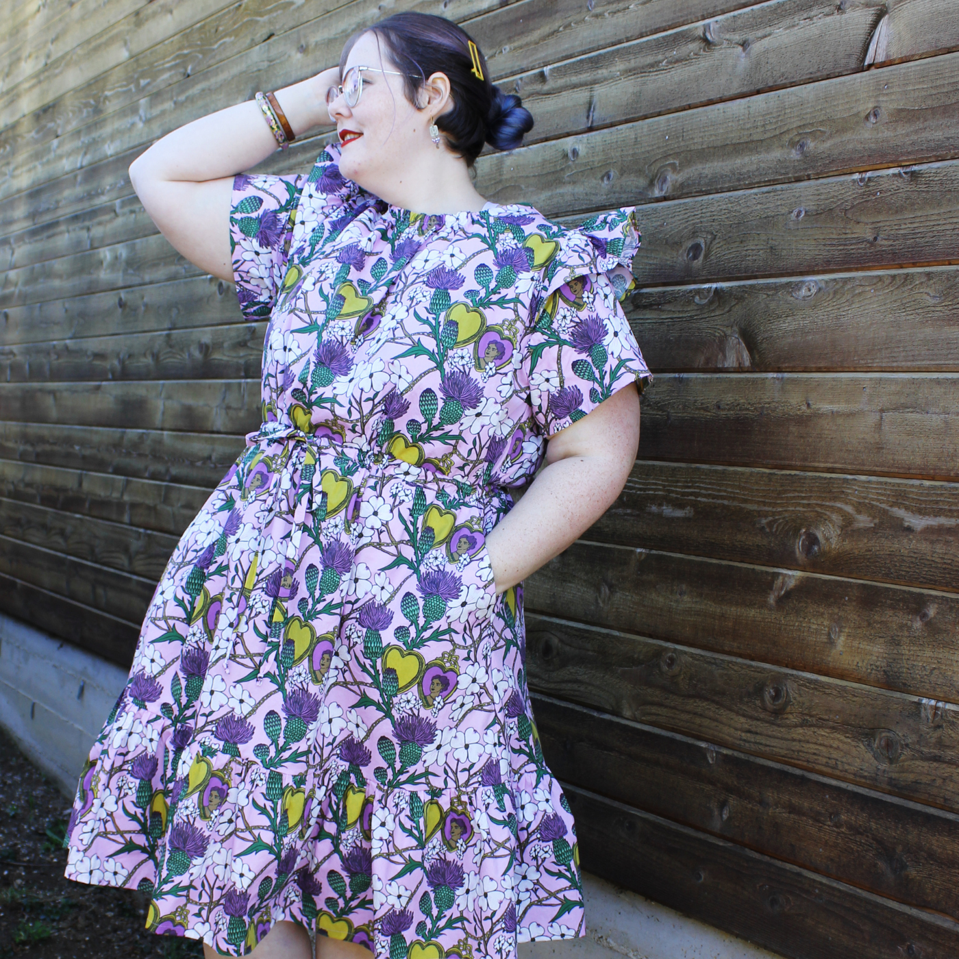Caroline stands in front of a wood-planked wall looking away from the camera with her left hand in a pocket and her right hand up against her head. She is wearing a dress with a lavender background, yellow hearts, purple and green thistles and small white dogwood flowers.