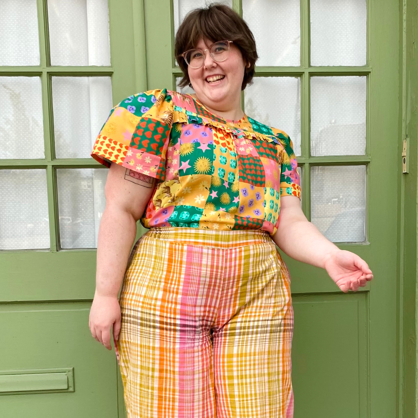 Caroline stands looking at the camera smiling in front of two green doors, the top half of which has rows of small rectangular windows. White curtains peek through the windows. Caroline wears a short-sleeved shirt with a patchwork print with small square patches of a number of designs, including red hearts on a green background, purple flowers floating on an orange background, pink stars and orange circles on a pink background and more. She also wears orange, yellow, pink and white plaid pants. Her left hand is held up and a little bit in front and her right hand is down by her side.