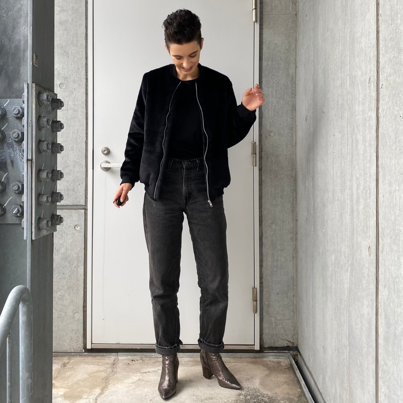 Emilia stands at the end of a concrete walkway with a gray wall to the right and a gray door and wall behind them. They are wearing black jeans, a black shirt and a black unzipped zippered jacket and black short heels. They are looking down and smiling, their left hand is up by their left shoulder and their right hand is down by their waist.