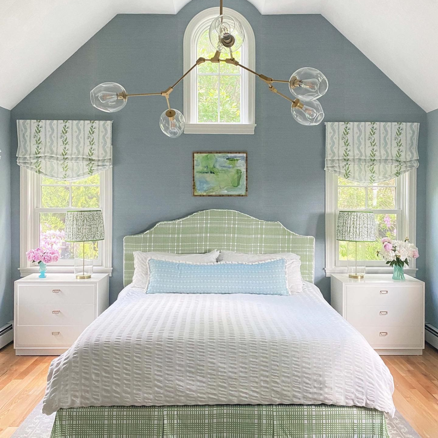 A full-sized bed is in the middle of a bedroom with rectangular windows on either side and a smaller window with a rounded top above it. The headboard and the bedskirt are both a green and white plaid. The bedspread is white and textured and two white and textured pillows are at the top of the bed behind a blue lumbar pillow with white design accents and fringe. A large bronze chandelier is above the bed with long extensions leading to globe ovals with lightbulbs. White bedside tables with three drawers are on either side of the bed. They both have vases with flowers in them (on the left a blue vase and pink flowers, on the right a green vase with pink and white flowers) and a gray-and-white lamp. White valances with alternating rows of blue wavy lines and sprigs of green leaves are on each of the rectangular windows.