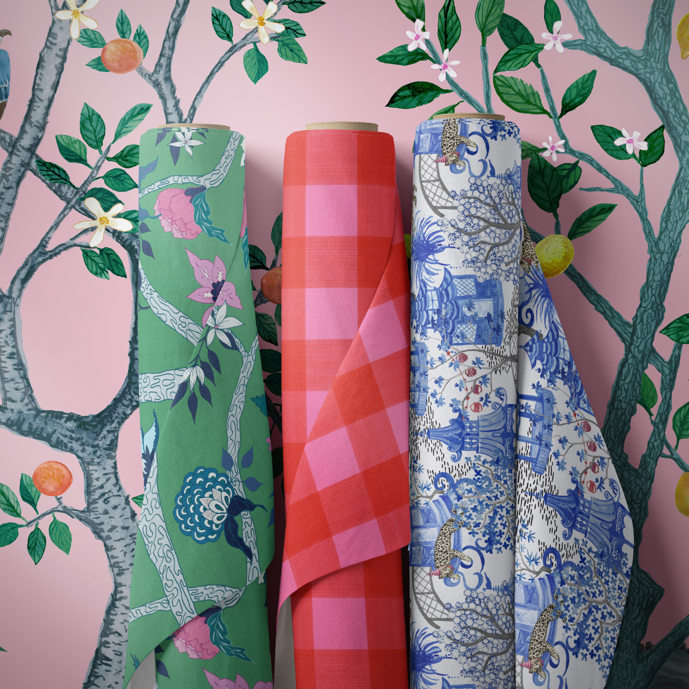 Pink wallpaper with wispy trees with oranges and small white blooms is behind three spools of fabric. The fabric on the left has a green background and white trees with blue accents with blue-and-white blooms as well as pink blooms and blue leaves. The middle fabric is a large pink-and-red-checkered design. The design on the right features repeating blue pagodas amidst trees with lots of small round white flowers, small gray fences and black-and-white leopards sleeping on tables and walking around the pagodas.