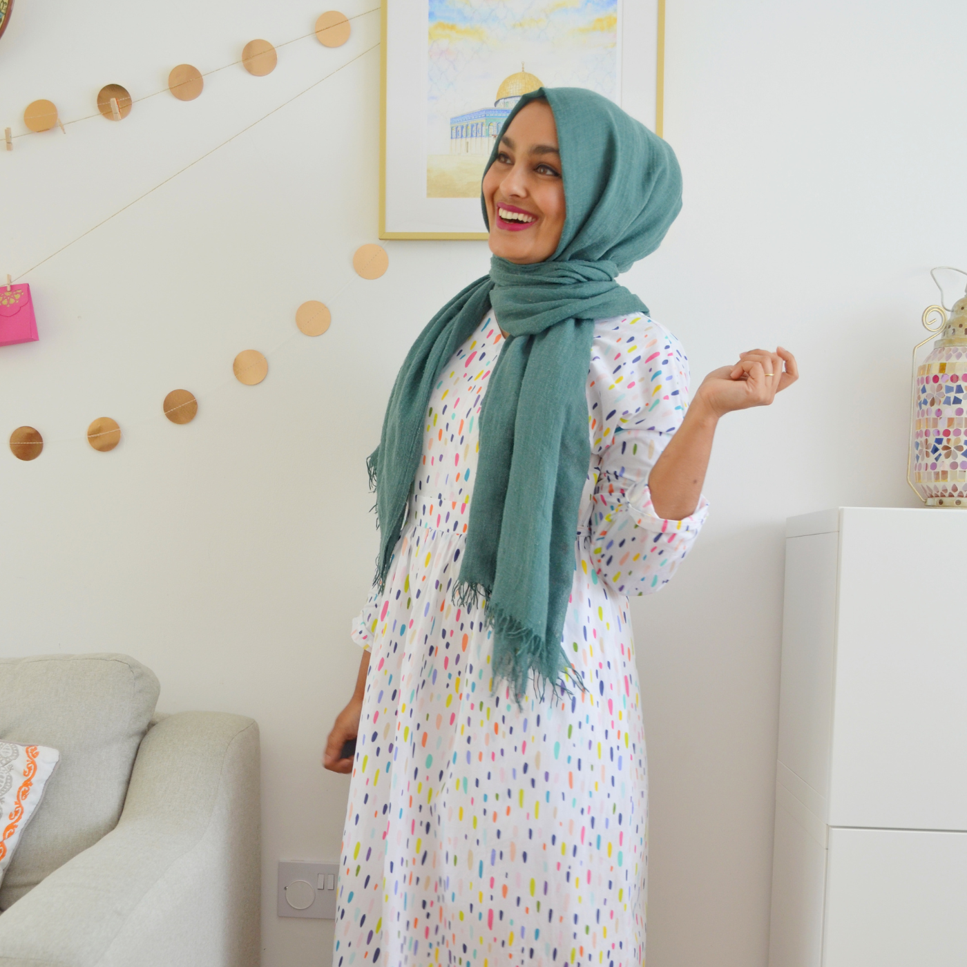 Rumana Dawood stands smiling, looking up and away from the camera. She is wearing a light blue hijab and a long-sleeved white dress with small blue, pink, olive green and dark blue brushstrokes. She is standing in front of a blue-and-yellow painting and next to a white chair on the left and a white bookshelf to the right. A garland of light brown dots hangs over the chair and from the painting, extending to the left.