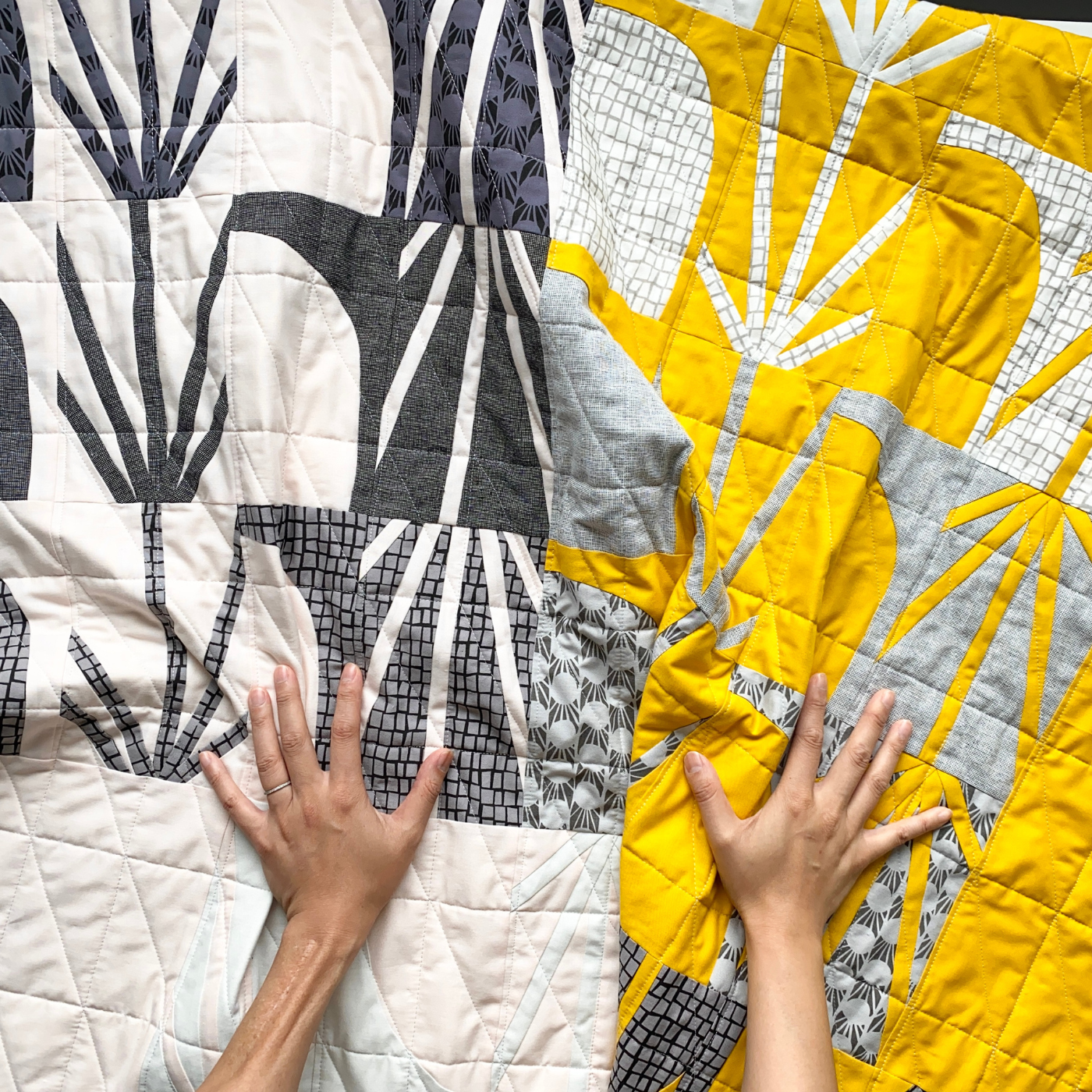 Two hands and forearms reach out and touch a quilt that is all white in the bottom left; white, black and gray geometric designs in the top left; yellow and white geometric designs on the top right; and yellow, gray and black geometric designs on the bottom right.