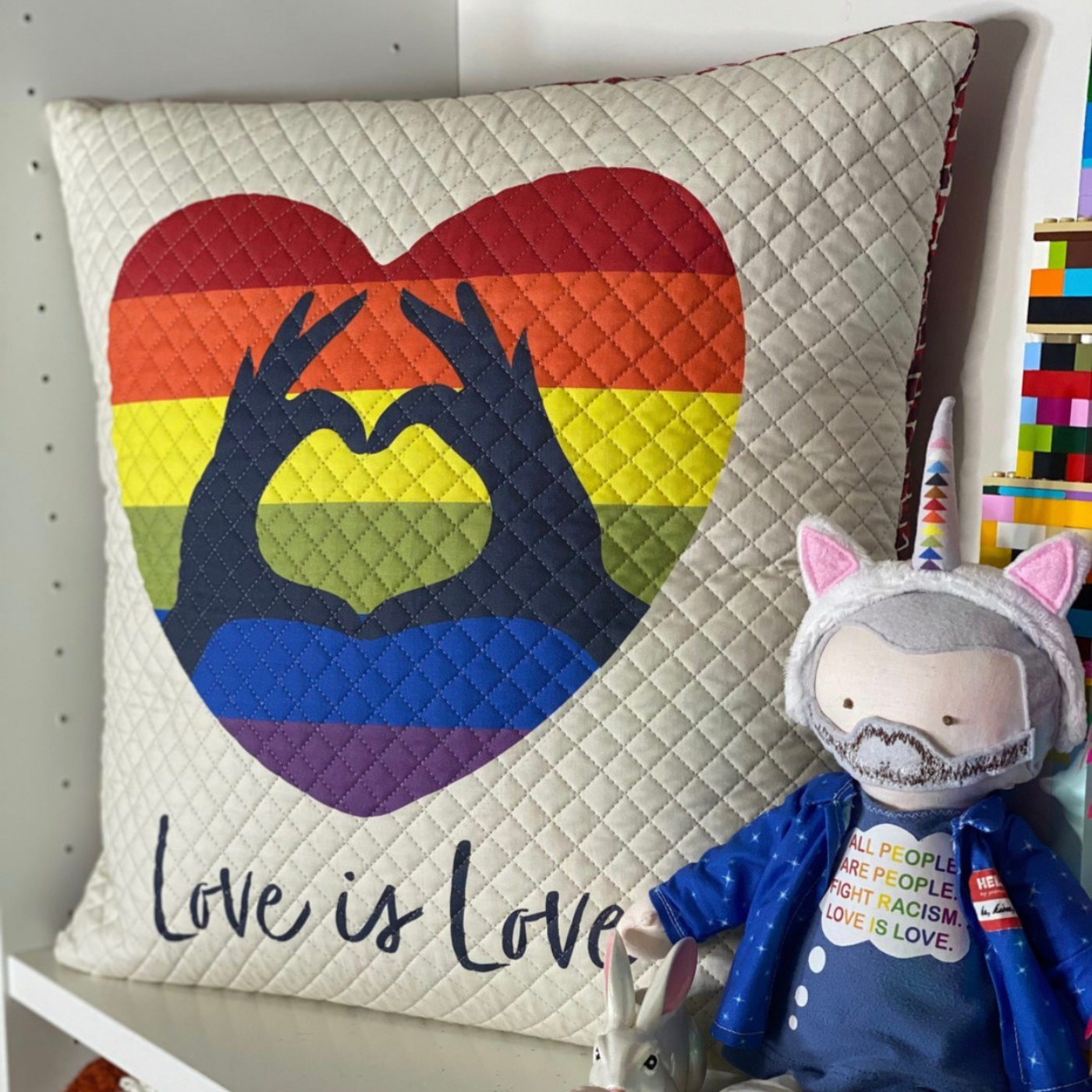 A cream quilted pillow with a rainbow striped heart in the middle, showing two black hands held together forming the shape of a heart, and the words “Love is Love” in black cursive font underneath, is on a white bookshelf. Next to the pillow is a small gray plastic rabbit and a small sofitie of a person with gray hair and a gray beard, wearing a blue hoodie and a blue shirt that says “All people are people. Fight racism. Love is love.” in rainbow letters, and a headband with pink cat ears and a rainbow unicorn horn.