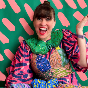 Katie Kortman sits against a green wall with short salmon-colored lines in repeated rows behind her. Her left hand is up in the air and her right hand is down in her lap holding a fabric stack of Katie’s designs that spills toward the camera. The designs are a range of florals and solid backgrounds with short repeated lines in contrasting colors. Katie wears a dress made out of several of her brightly colored floral fabrics with a green detachable collar.