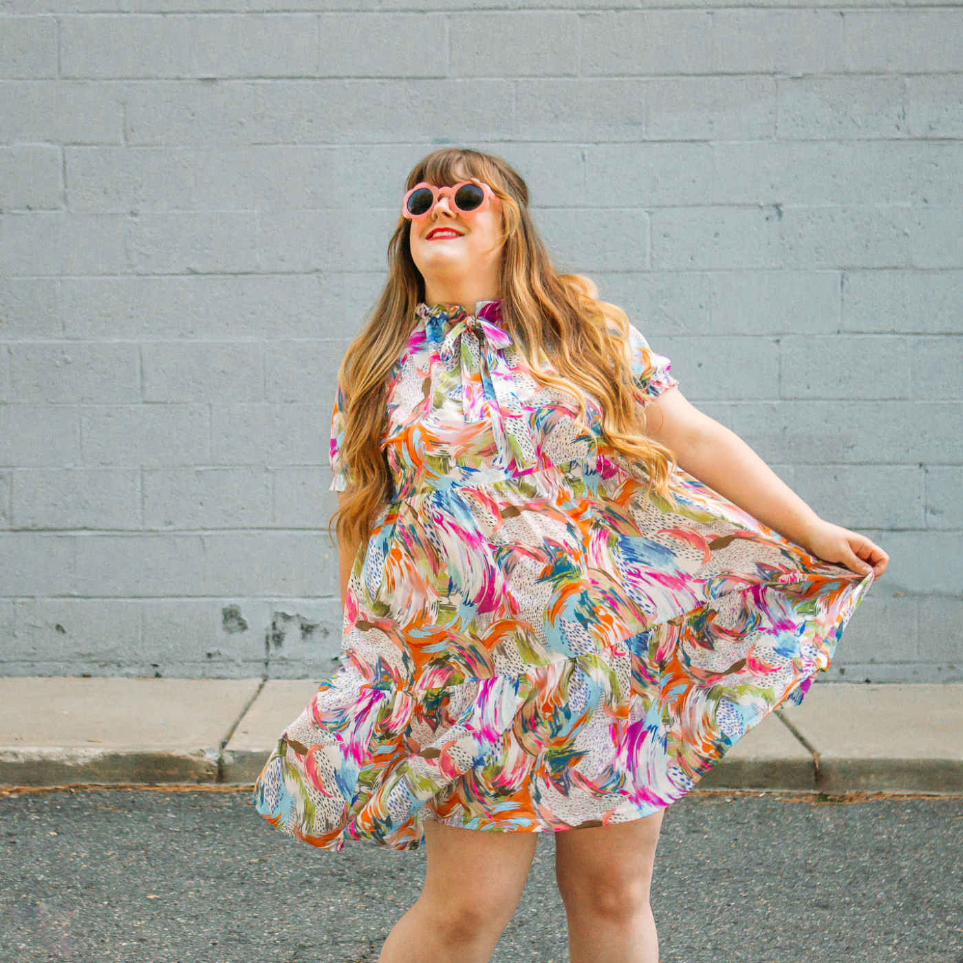 Meg Fleshman is looking up towards the sky and smiling. She is wearing round pink sunglasses and a flowing white knee-length short-sleeved dress with a design with rainbow brushstrokes grouped together in arcs around groups of smaller black brushstrokes. Her left hand is holding out the dress to the side. Behind her is a light blue concrete wall.