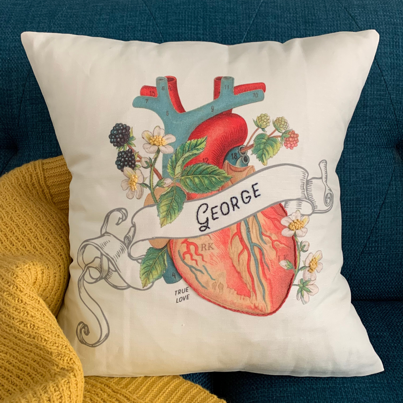A cream throw pillow with an anatomical heart design printed on it has a banner running through the middle of the heart that says “George,” which is stitched in black font. The pillow sits on a blue chair next to a yellow blanket.