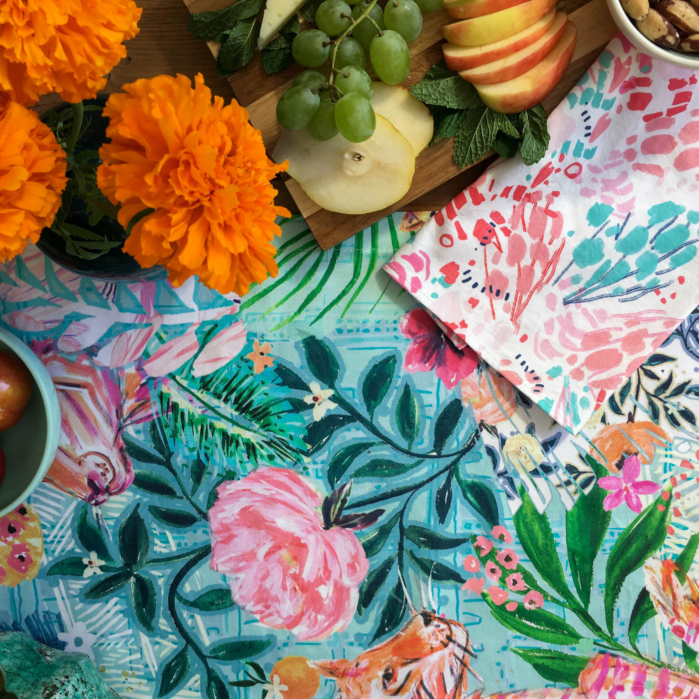 A cloth napkin with a white background and pink, red and turquoise brushstroke flowers sits next to a vase of small orange flowers and the edge of a wooden cutting board with mint leaves, apple slices, grapes and a small white ceramic bowl of Brazil nuts. On the table is a tablecloth with a turquoise background and a design featuring large orange tigers surrounded by large pink blooms, small oranges and sprigs of green leaves.