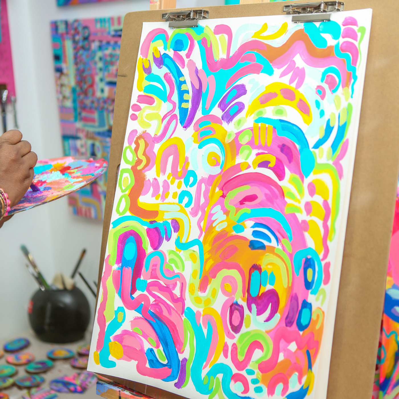 A hand partially shown in the middle lefthand side of the photo dips a paintbrush in a bit of purple paint on a full color palette. A piece of paper on an easel to the right is full of geometric squiggles in hot pink, light pink, orange, turquoise, dark blue and more. A similar painting is on the wall to the left of the easel. Small round colorful paintings are on the ground to the left.