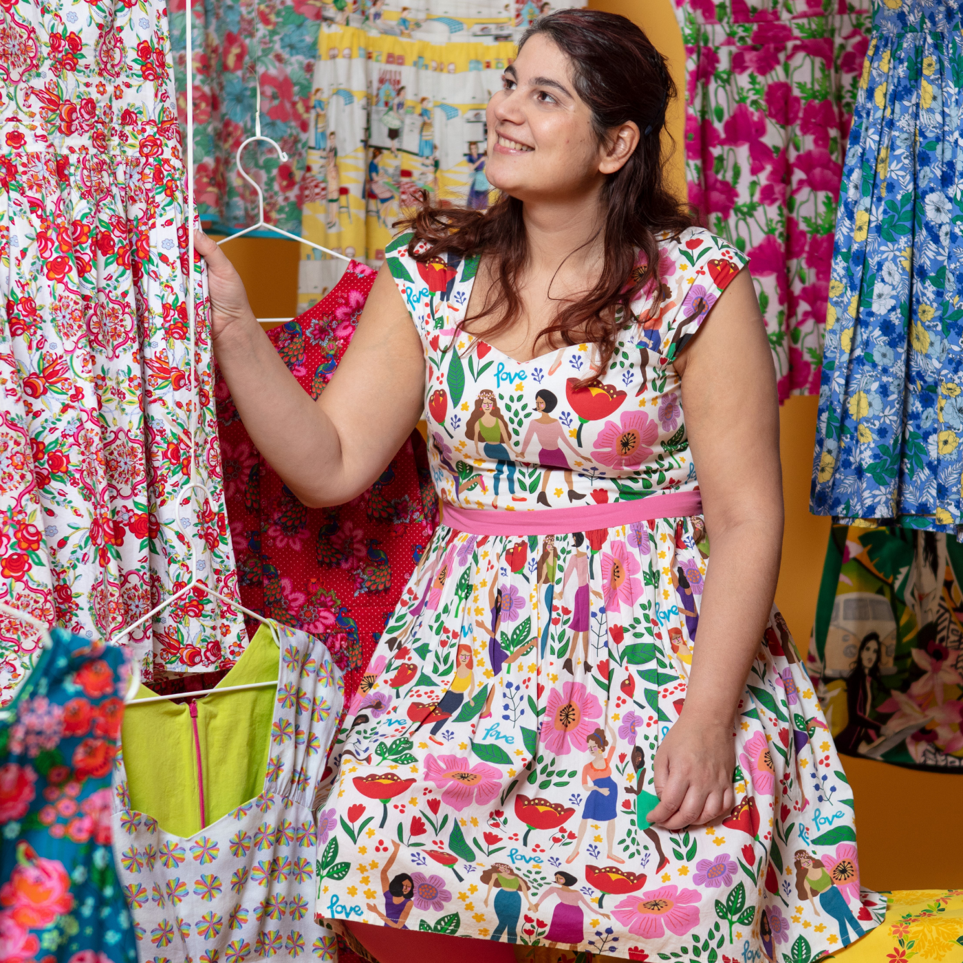 Sangeeta sits surrounded by colorful dresses and wears a dress with women holding hands, doing yoga and walking through large pink, red and purple flowers. She is looking up and away from the camera and smiling with her right hand on a white-and-red floral print dress.