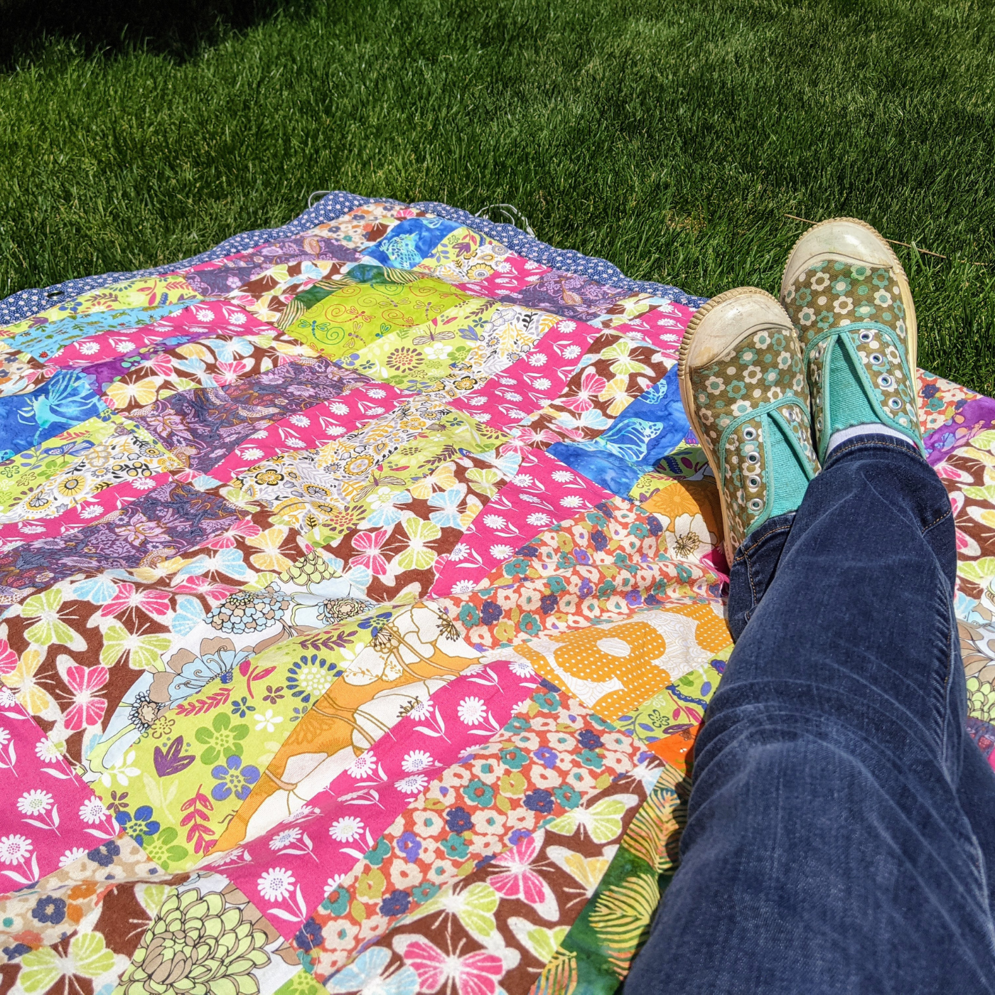 A colorful patchwork blanket rests on a bed of grass. Two legs extend into the lower right handside of the blanket, wearing jeans and green-and-blue floral print shoes.