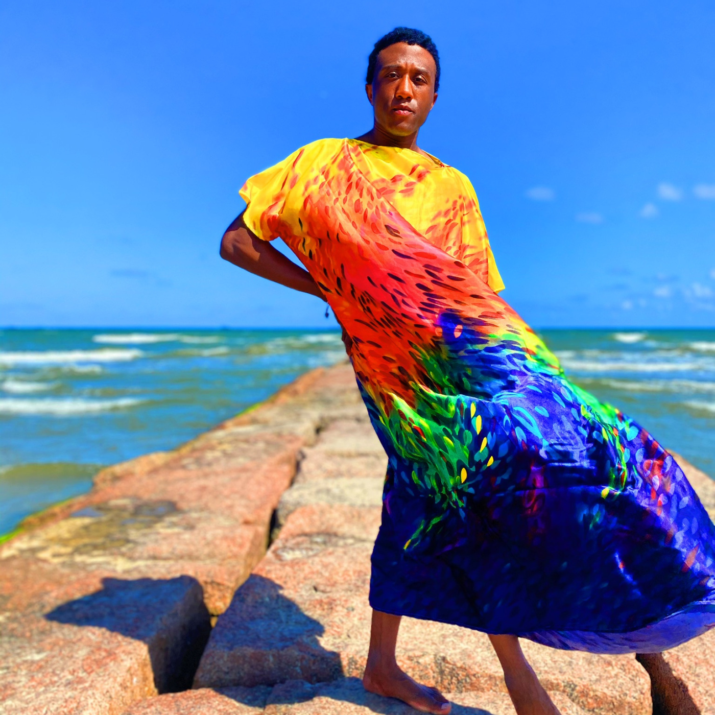 Terrance stands on a beach and looking at the camera, the ocean is behind him. He is wearing a yellow, red, green and blue caftan that is blowing in the wind to the right. His right hand is at his hip.