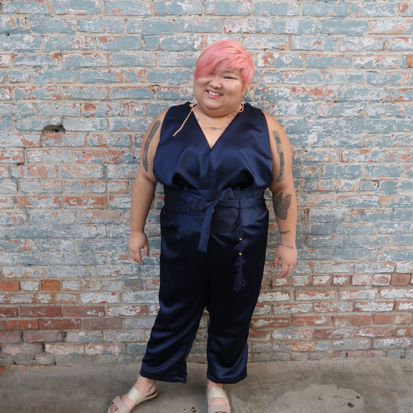 Teukie stands against a red brick wall painted gray, which is chipped and fading. They are smiling at the camera and wearing a shiny navy jumpsuit and gray sandals. Their arms are at their sides.