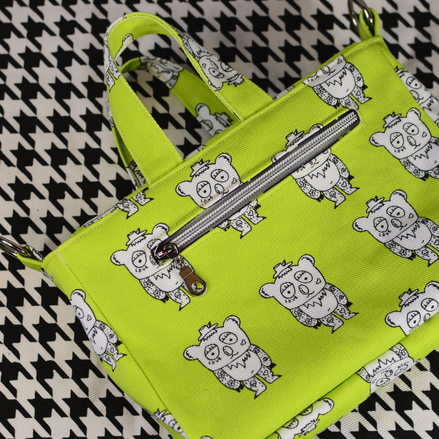 A small neon green bag with two handles and a zippered section in the front featuring rows of white bears with tattoos wearing hats outlined in black. The bag is lying on a black-and-white geometric print surface.
