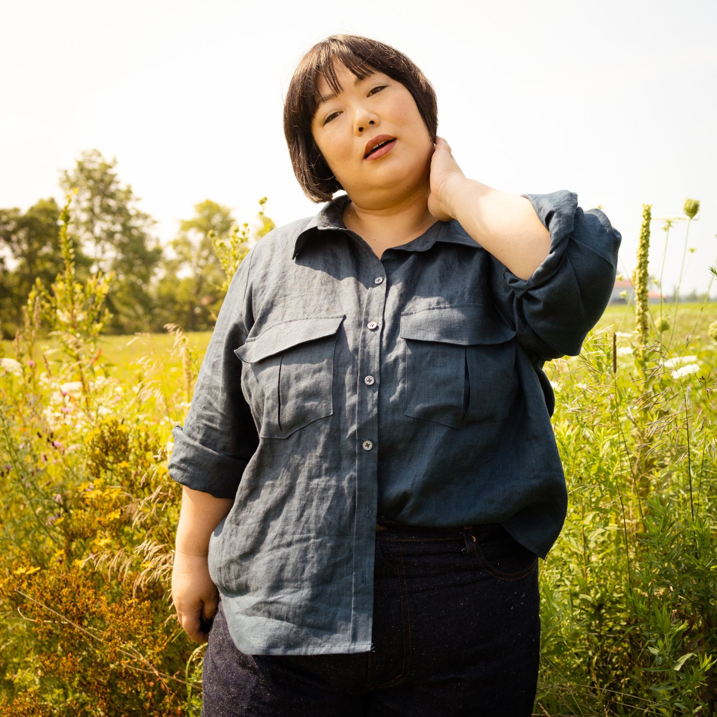 Leila stands in a field around tall small white flowers. She is holding her right hand up to her face and wears a long-sleeved button up shirt with the sleeves rolled up and black pants.
