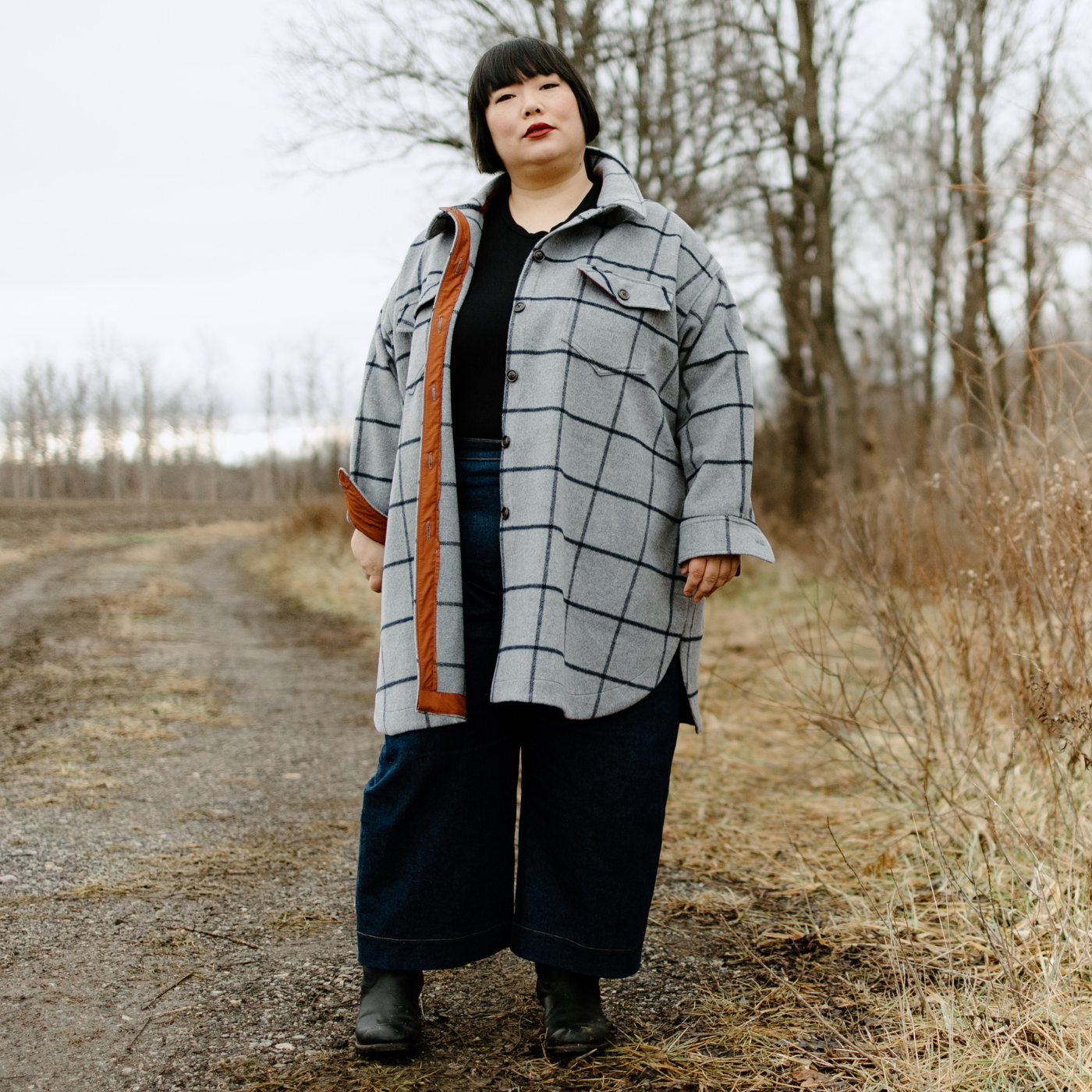 Leila stands looking at the camera and at the edge of a gravel road. She is wearing a gray coat unbuttoned with vertical and horizontal black stripes. The inside button band of the jacket is dark orange. Underneath the jacket she is wearing a black shirt and dark blue wide-leg pants.