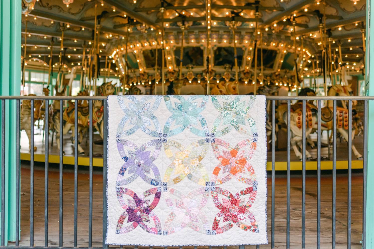 A quilt of large interlocking four-pronged stars, each a different color, ranging from light blue to dark rad on a white background. The quilt is hanging on a black metal fence outside of a lit-up carousel inside a pavilion.