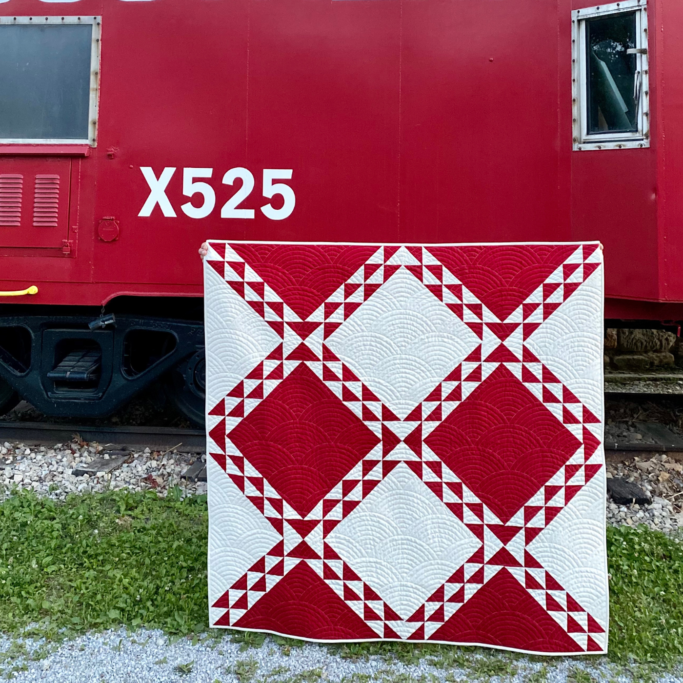 A red-and-white quilt featuring alternating rows of large red and white diamonds outlined by two small rows of smaller red and white alternating diamonds is in front of the side of red train car that has two small square windows, one on the left, one of the right that says X525. The quilt is standing up right in front of the train car on a patch of gravel and grass.