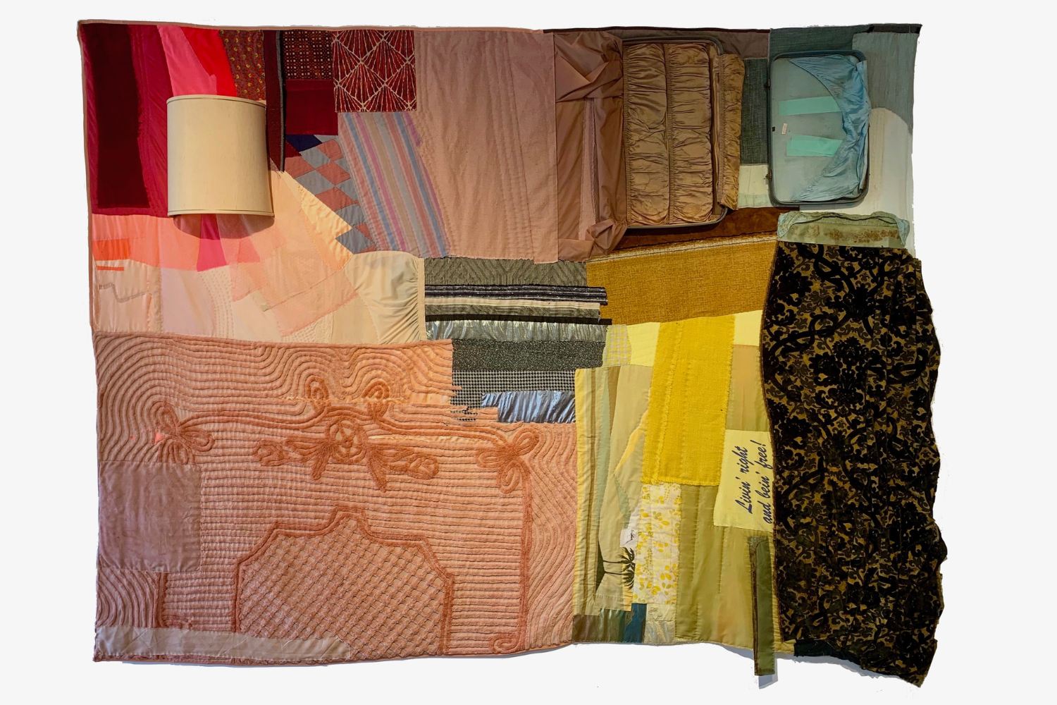 A hand-stitched quilt of many quilted blocks, each featuring items you might find in a room in a home, an orange-and-white bedspread, several strips close together in varying shades of grey to resemble a staticky television screen and more. Along with the blocks there are several 3-D elements, such as a white lampshade, part of which is sewn directly into the piece, and an actual small vintage blue suitcase.
