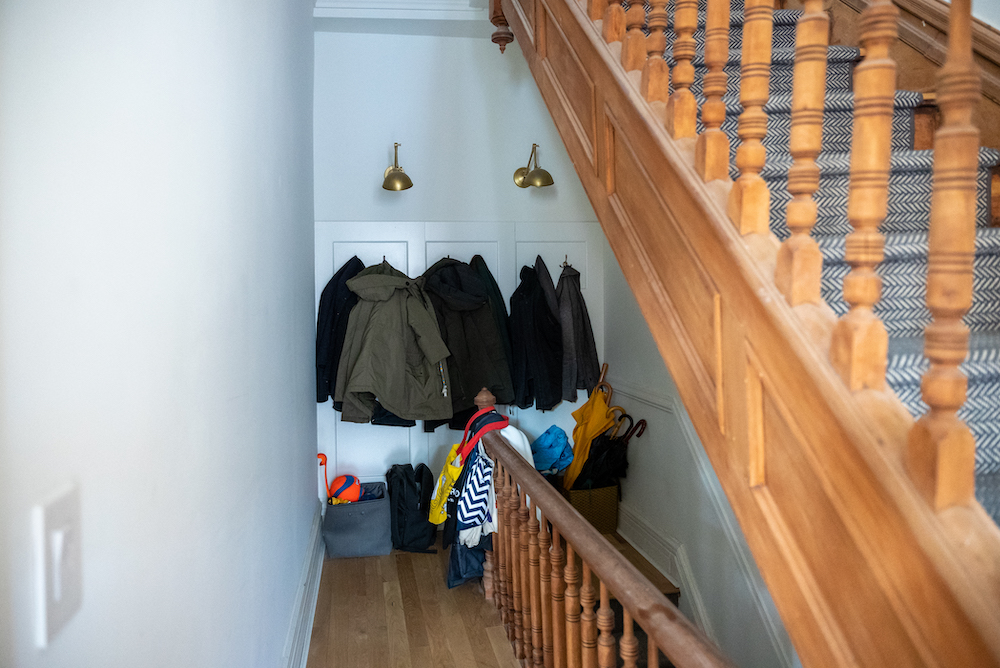 hallway at the top of a staircase surrounded by white walls. White, wooden coat panel and hooks with black and dark-colored coats hanging on the hooks. Baskets and umbrellas on the floor beneath the coats. Brass sconce-lighting above the coats.