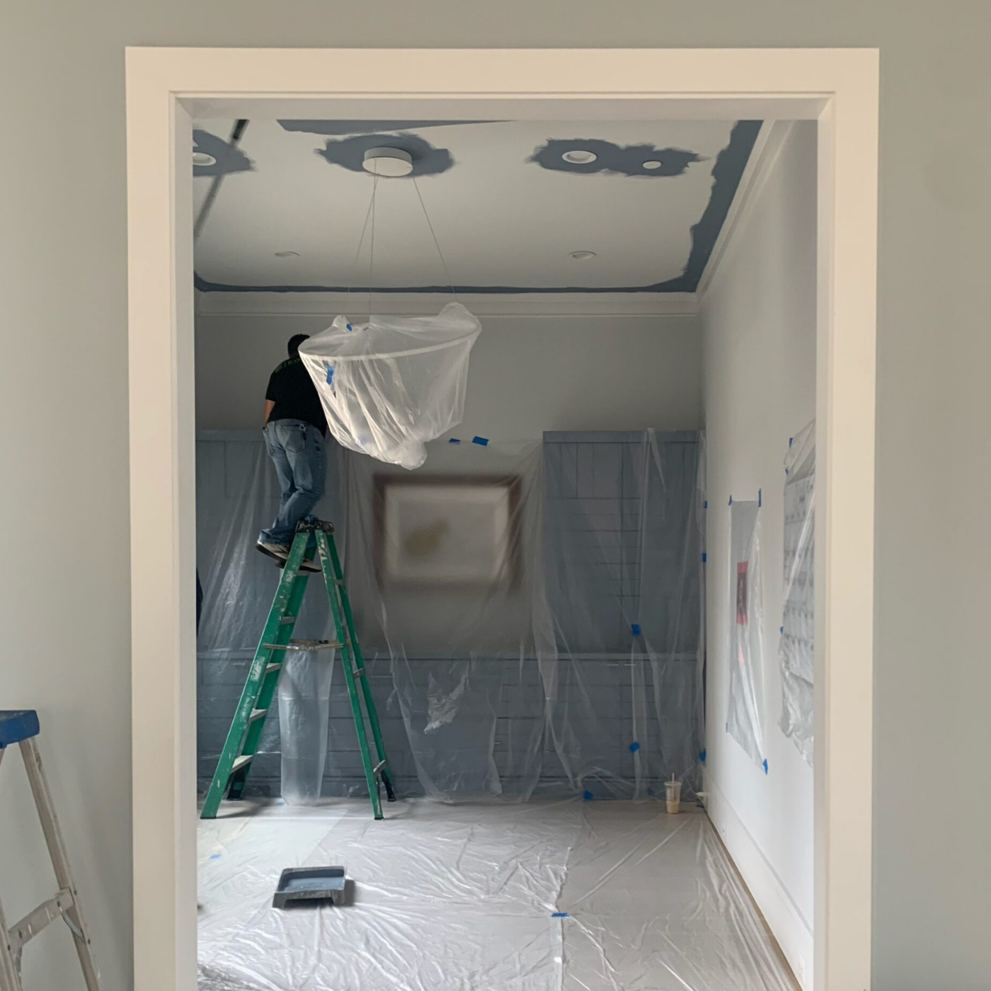 Looking into a home office from a hallway, a white door frame and gray walls frame the view. Part of a ladder is against the leftside outer wall. A large plastic sheet covers built-in gray cabinets that are taller on the left and right sides and table height in the middle. A rectangular piece of art hangs over the middle section and has a brown frame, white mat and a small darker design rendered too blurry to discern under the plastic. A person stands in front of the cabinets on the highest step of a green ladder and is facing the back wall. The white carpet is also covered in plastic and there is a paint tray full of gray-blue paint in the middle of the floor. The ceiling is partially painted, with gray-blue paint around three recessed lighting fixtures and a fixture holding a large white ring light in place that hangs from the ceiling.