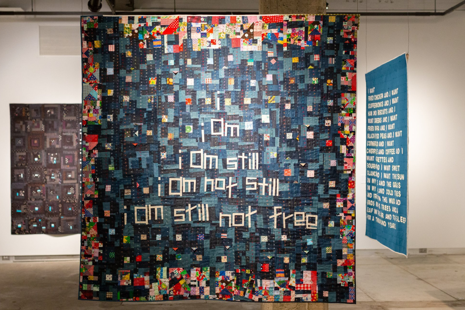 Photo of a quilt displayed from the ceiling in a gallery. A blue quilt with white all caps text is to the right and a quilt with a black background and rows of small patchwork squares in mostly dark fabric is behind and to the left. The main quilt has lowercase text in white fabric that reads “i am / i am still / i am not still / i am still not free.” Behind that text are small blocks made out of fabric that is black and various shades of blue with a few brightly colored small blocks peppered throughout. The border of the quilt is comprised of small brightly colored blocks worked together to form a border.