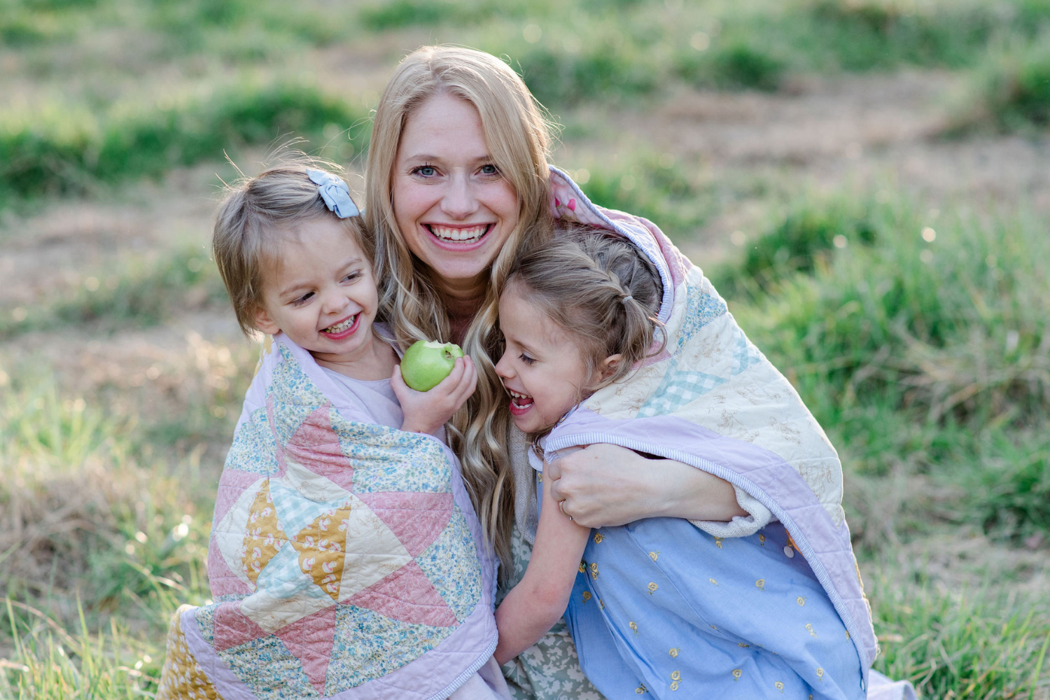 Michelle Collins and her two daughters are smiling and wrapped in a quilt comprised of small triangle blocks, some are blue and yellow floral, some are pink, some are white. The quilt has a thick lavender border. They are in a grassy field.