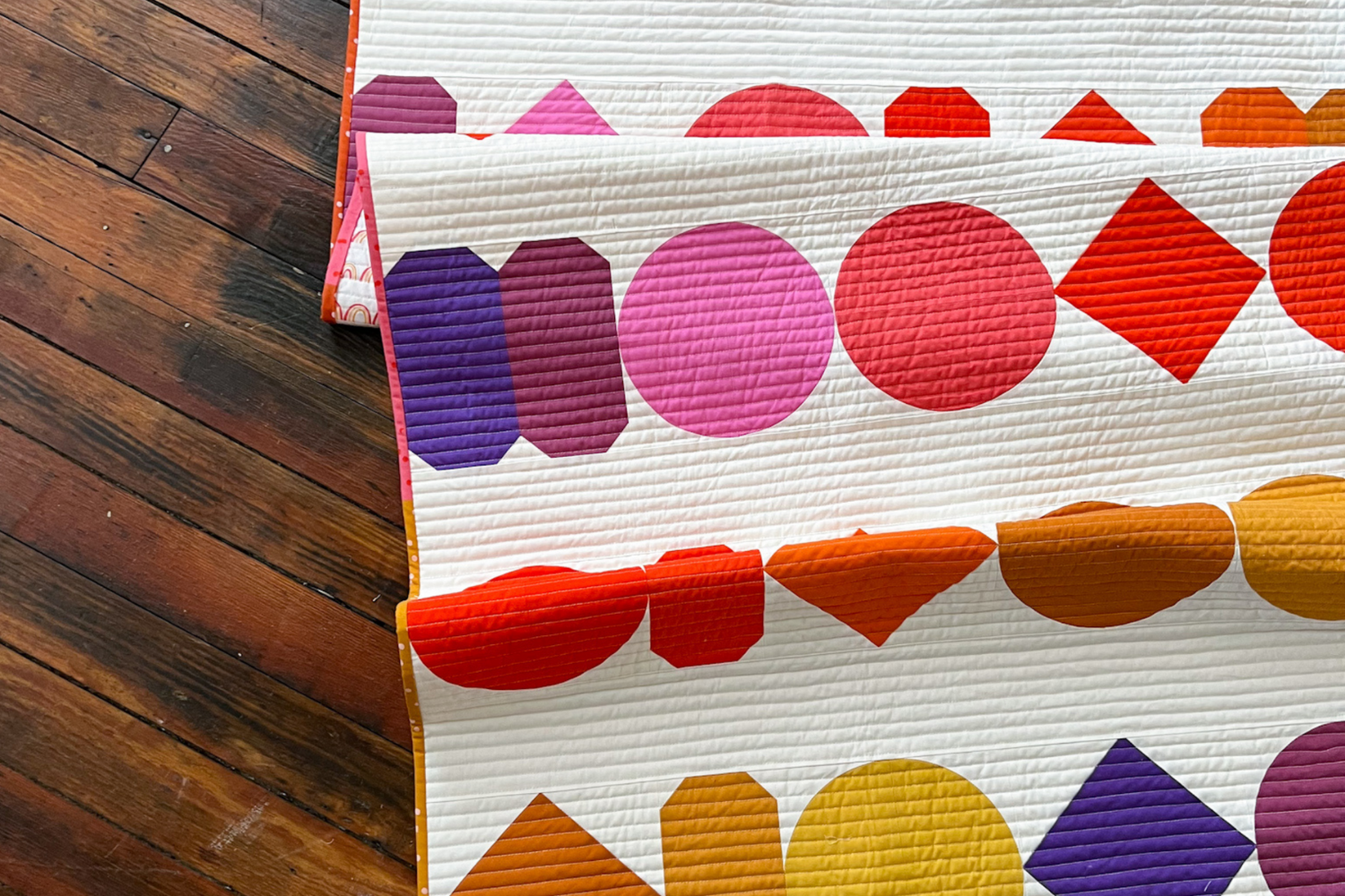 A quilt lies on a dark wooden plank floor. It has a white background and rows of bead shapes, round, oval, square, and more, in jewel tone hues, pink, red, purple and yellow.