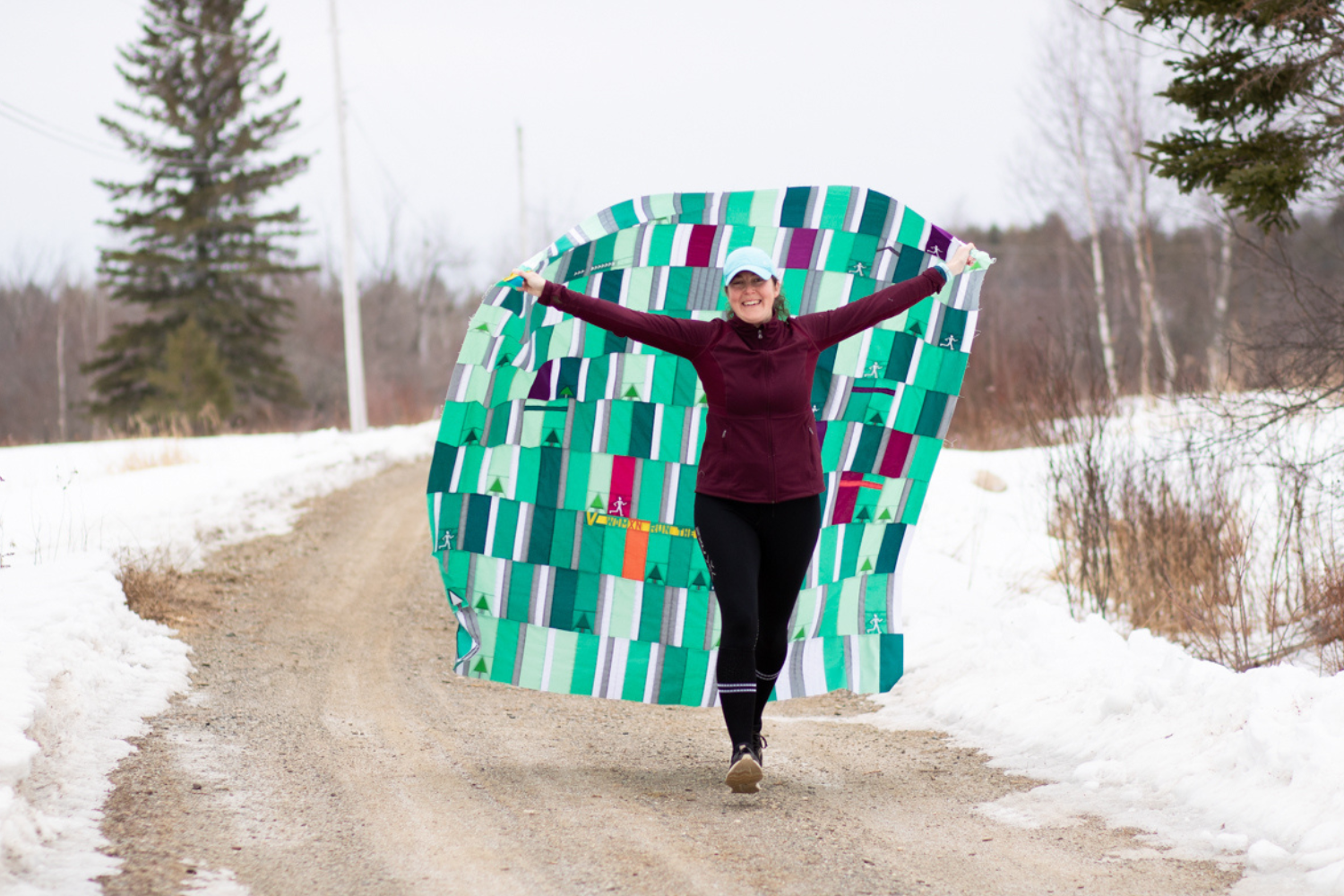 Kitty smiles and holds up her My 2020 Miles Quilt behind her as she walks down a gravel road with snow on either side of the road. A gray sky and bare brown trees, and one tall green fir tree, are behind her. Kitty is wearing black pants, a burgundy long-sleeved top and a mint green baseball cap. The quilt is comprised of small mostly green blocks (in both dark green and shades of lighter green), with a few white, red and orange blocks.