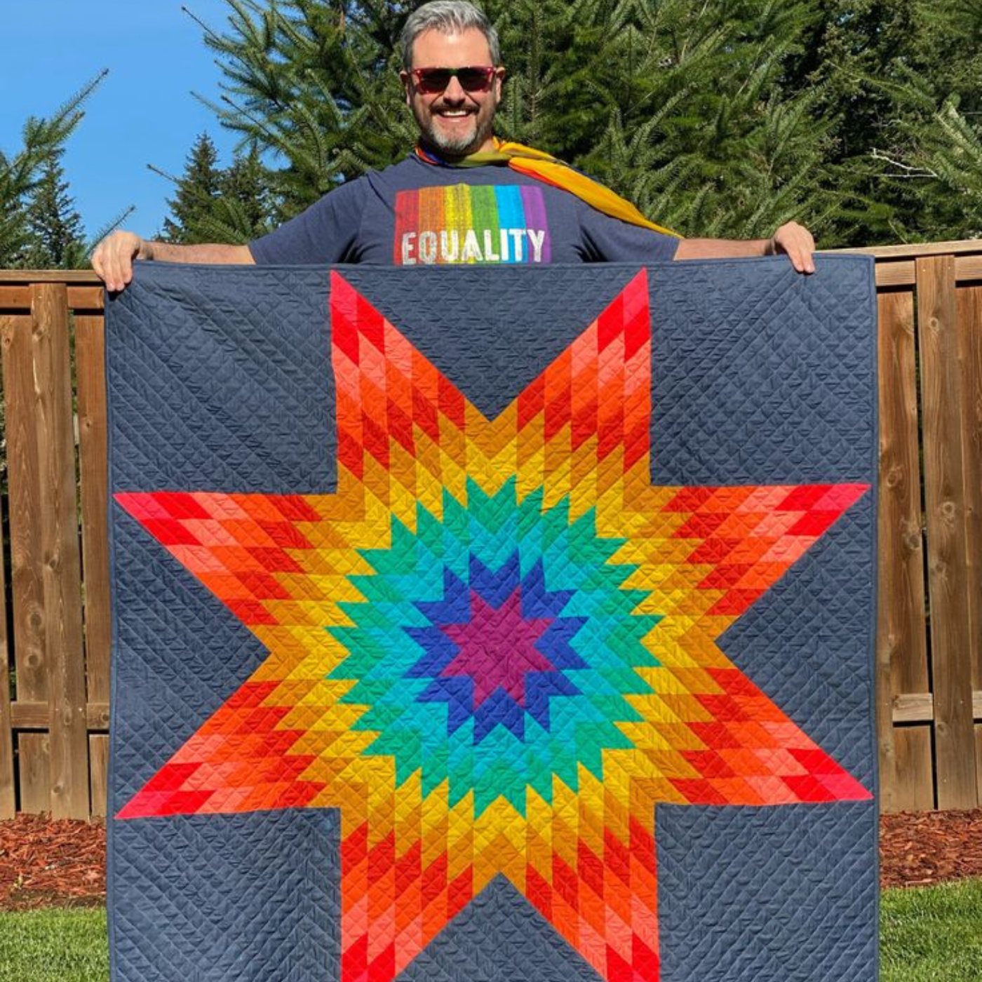 Mathew is looking at the camera and smiling, holding up a quilt with a gray background and a large rainbow star design on it. The center of the quilt is purple, then extending outward, blue, teal, green, yellow, orange and red. He is wearing sunglasses and a gray shirt with vertical rainbow stripes that says “EQUALITY” in all caps.