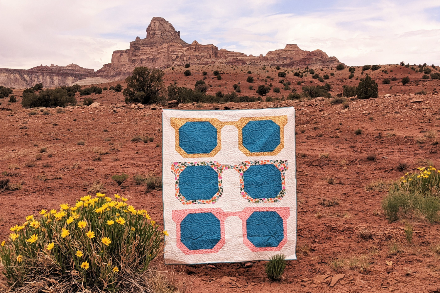 A quilt is held up in the middle of a desert. A large brown and tan rock formation is behind the quilt, slightly to the left side of the photo. Small green shrubs dot the brownish-orange desert behind and around the quilt, and small yellow flowers are growing on green stems directly to the quilt’s front left and to the quilt’s right. The quilt has a white background and three large pairs of sunglasses, one on top of the other, making three rows. They all have blue lenses; the frame on the top pair is yellow with small white dots, the frame on the center pair is an orange, blue and brown leaf design and the frame of the bottom pair is pink with small red dots.