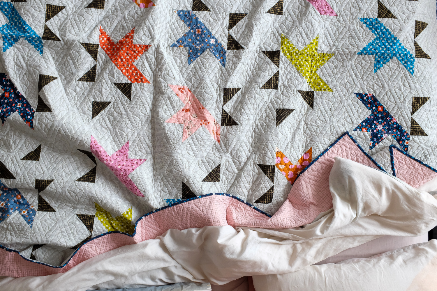 A quilt with small solid colorful slices of a starburst in reds, pinks, blues and yellows repeat next to smaller black triangles, leaving part of each star’s center in white, which is the same colors as the quilt’s background.