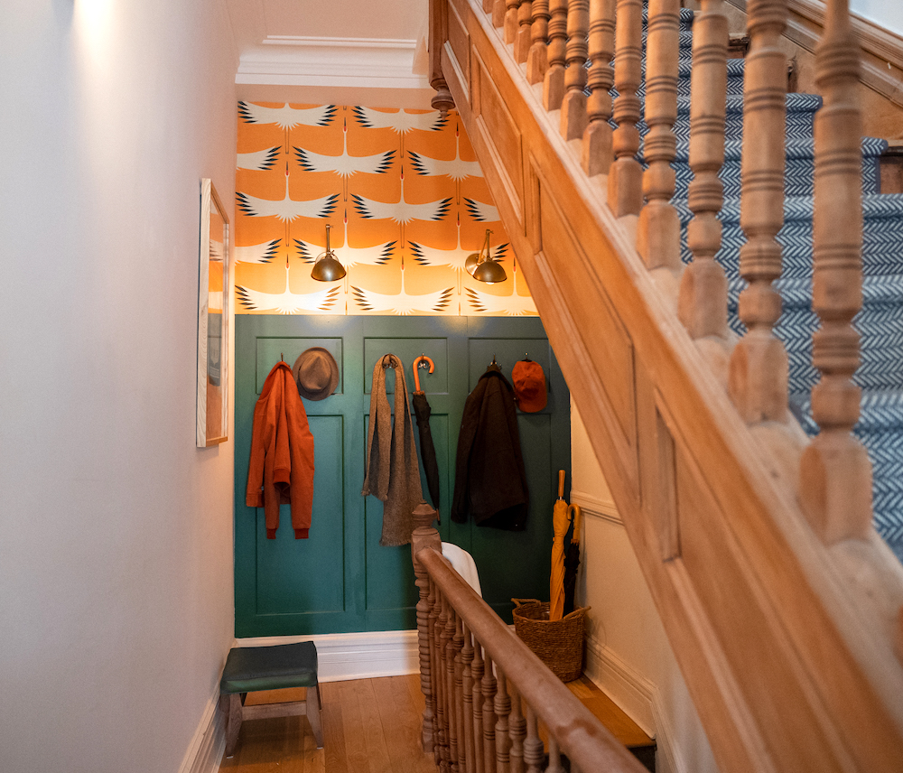 A hallway at the top of a stairway featuring dark yellow wallpaper with white cranes, and a dark green wooden coat hook panel. Coats and umbrellas hanging on the hooks.