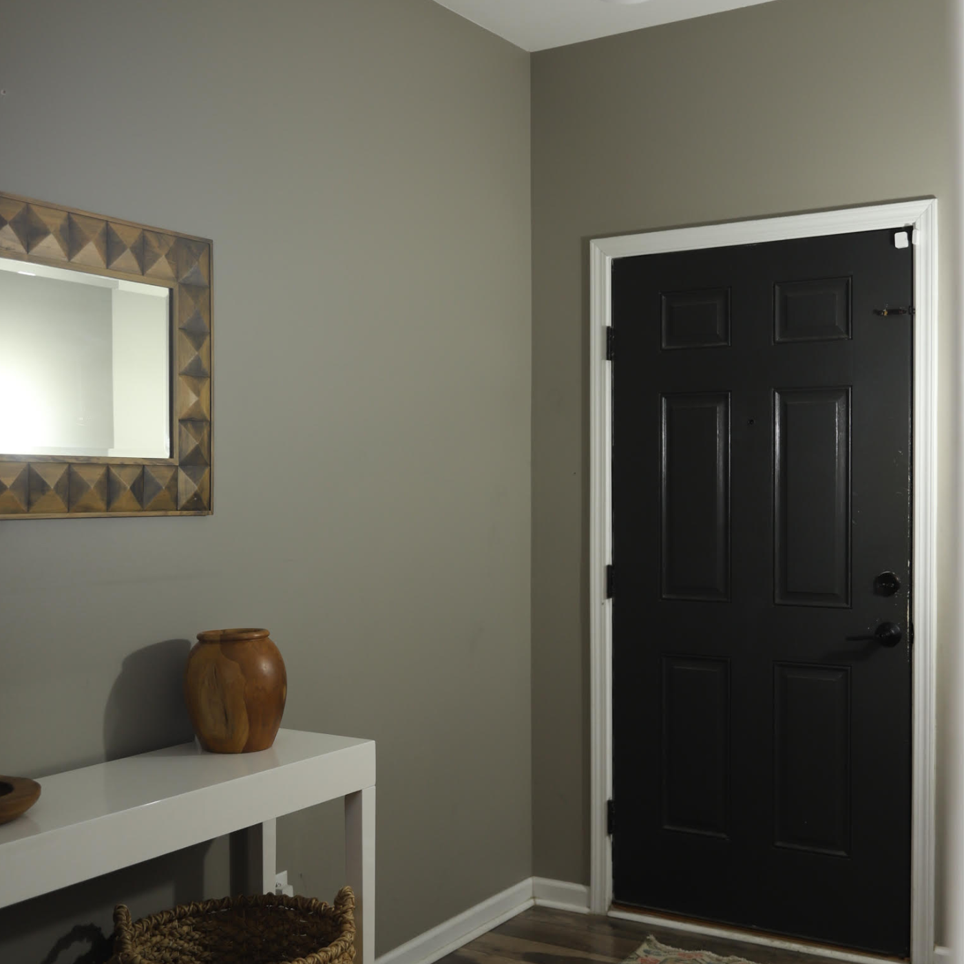 The entryway before the new grasscloth wallpaper glowup featuring gray walls, white trim and a black door. A rectangular mirror is placed lengthwise above a small white table with a wooden vase on it and grass baskets below.