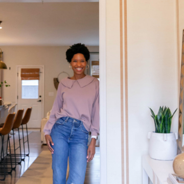 Tiffanie stands in the new entryway in her home and looks at the camera smiling. She is wearing a mauve blouse tucked into blue jeans and tan sandles. The design on the wallpaper has a cream background and two small dark tan repeating stripes. To the front left is a small white table with a tall green plant in a white vase, a tan vase and a large rectangular mirror balanced against the wall and two large grass baskets underneath the table. Behind Tiffanie is a banquette with three tall brown chairs with black wire bottoms, a cream carpet over dark brown wooden floors and a white door leading outside.