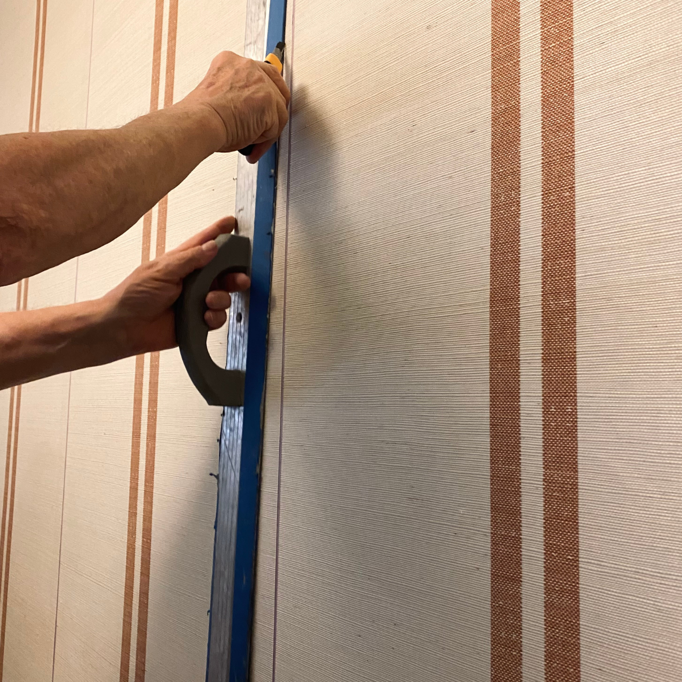 A photo of a wallpaper installation. A left hand holds a blue level while a right hand holds a small tool while installing wallpaper with a cream background and two repeating dark tan lines.
