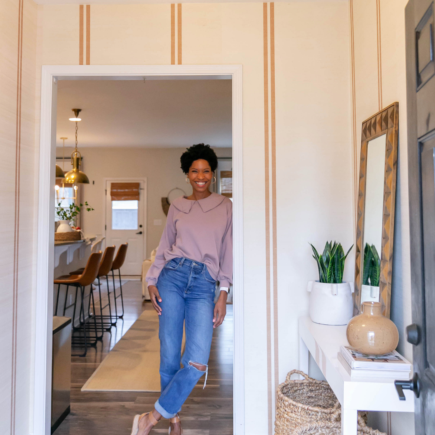 Tiffany stands in the new entryway in her home and looks at the camera smiling. She is wearing a mauve blouse tucked into blue jeans and tan sandles. The design on the wallpaper has a cream background and two small dark tan repeating stripes. To the front left is a small white table with a tall green plant in a white vase, a tan vase and a large rectangular mirror balanced against the wall and two large grass baskets underneath the table. Behind Tiffanie is a banquette with three tall brown chairs with black wire bottoms, a cream carpet over dark brown wooden floors and a white door leading outside.