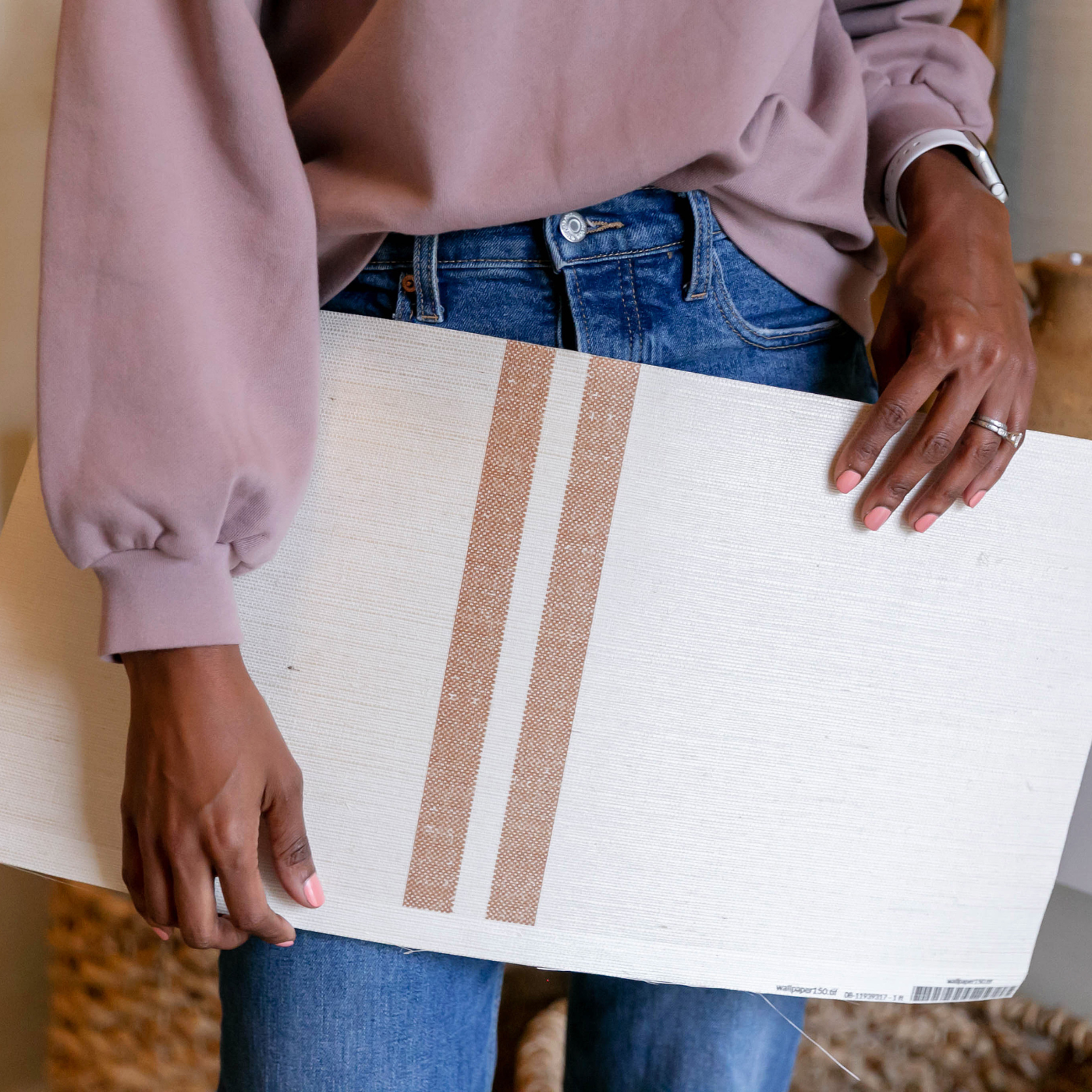 A photo of Tiffany holding a swatch of grasscloth wallpaper down by her waist. Her hands, torso and legs are shown. She is wearing a mauve blouse tucked into blue jeans. The design on the wallpaper has a cream background and two small dark tan repeating stripes.