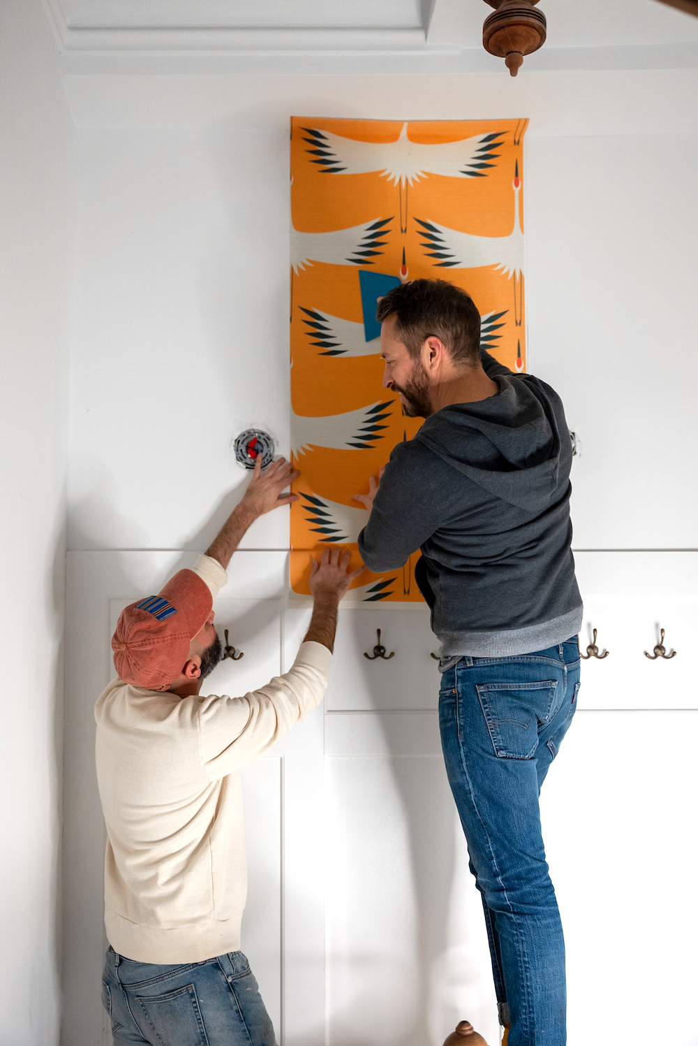 Two men working together to install dark yellow wallpaper with white cranes at the end of a white hallway. Wallpaper panel is being installed above an empty coat-hook shelf