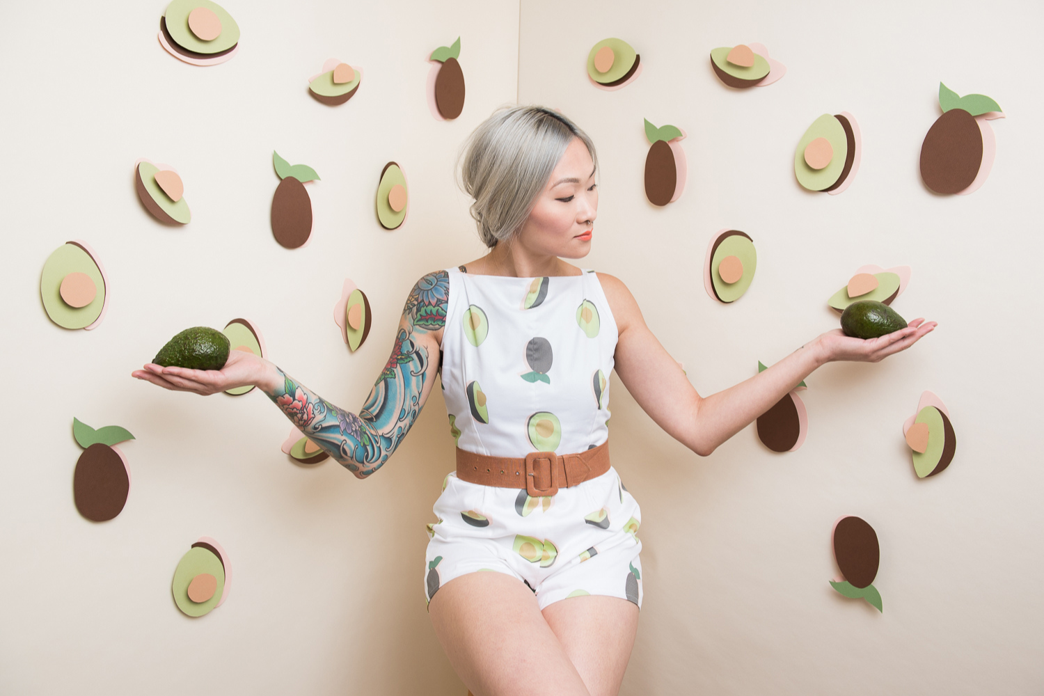 A model wears a romper with a white background featuring a design with both whole avocados as well as avocados cut in half showing their bright green pulp and brown pit. The model’s arms are both held out and each hand holds a whole avocado. The model is looking at the avocado on the left. Avocados constructed from paper, both whole and cut in half, are fixed to the walls behind the model to both the left and right. Caption: Model: Amy Vong. Photo: Kate Warren.