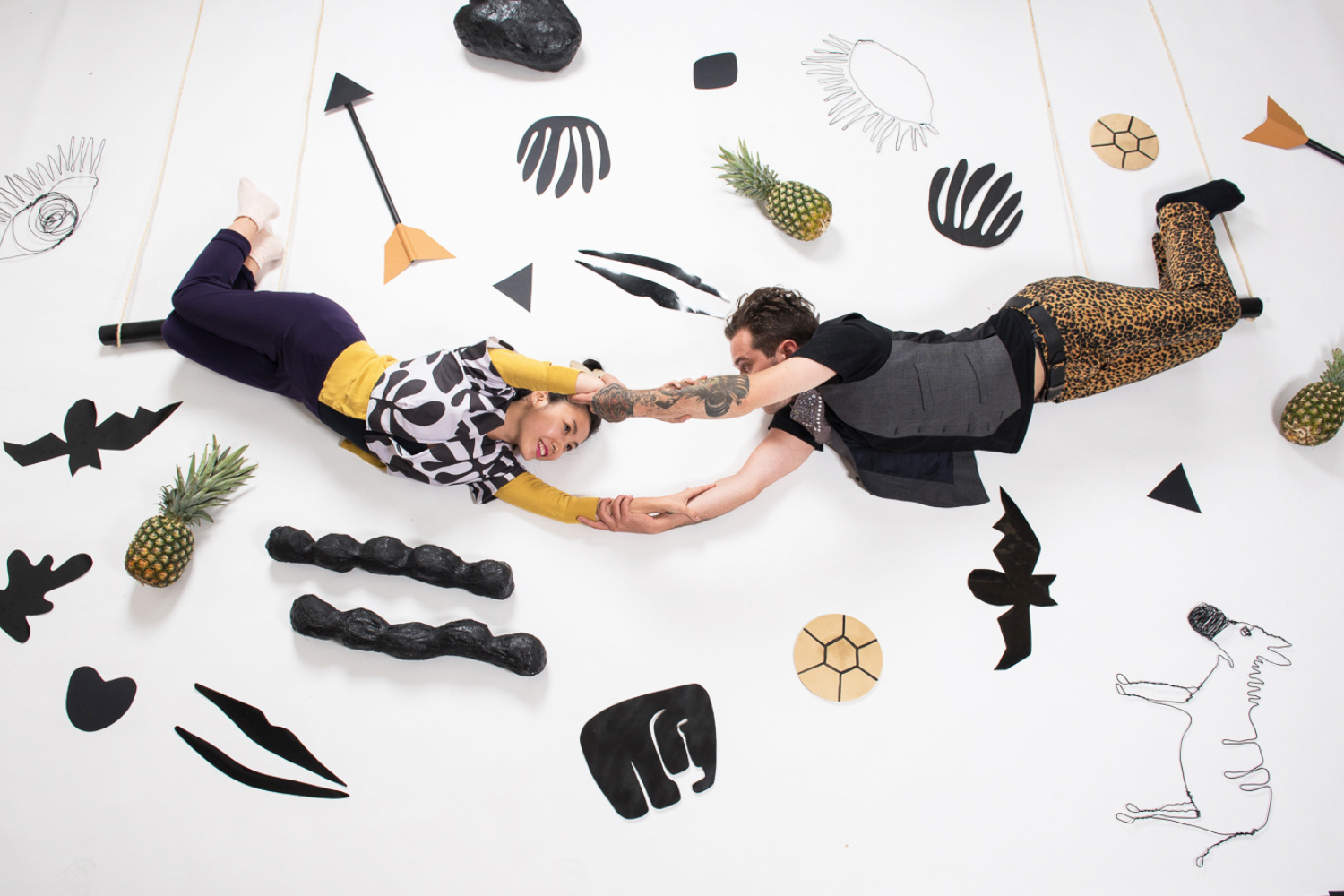 Two models grab hands as they hang from trapeze swings, one model is facing left and another model is facing right. A white wall is behind them. On the wall are a number of black geometric shapes, black bats, pineapples and small round shapes with black intersecting lines.